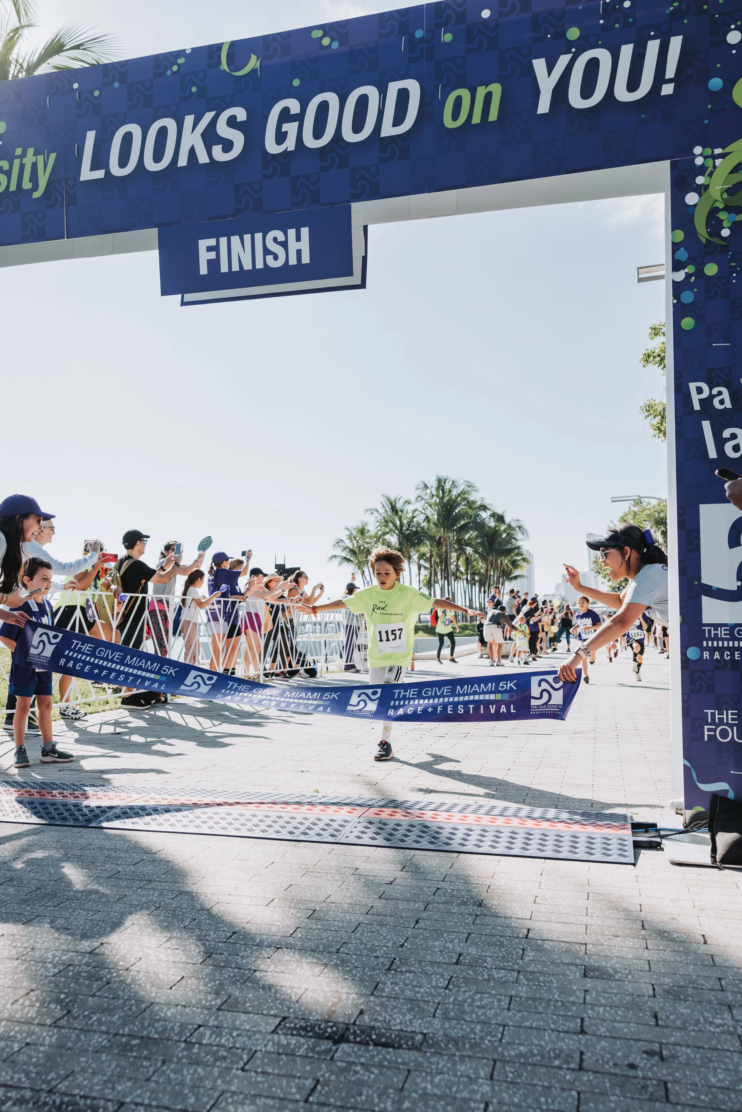 A young child crosses the finish line at 'The Give Miami 5K Race & Festival' event, with onlookers and supporters cheering on from the sides. The scene is set on a sunny day with palm trees in the background.