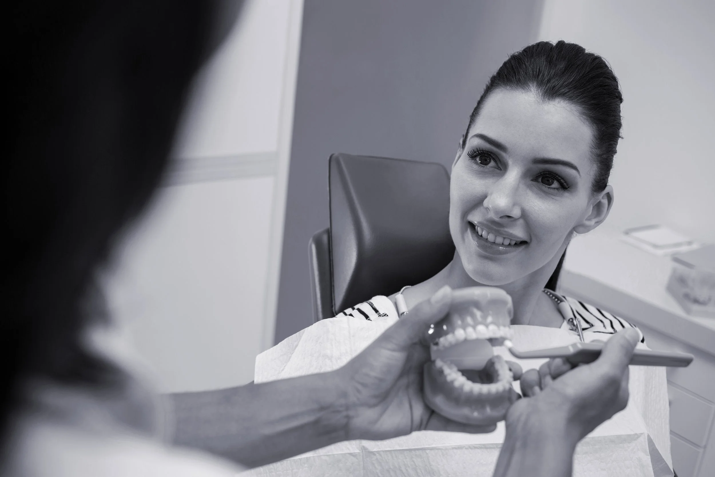 A woman at the dentist's office smiling while a dental professional holds a plastic model of a set of teeth.