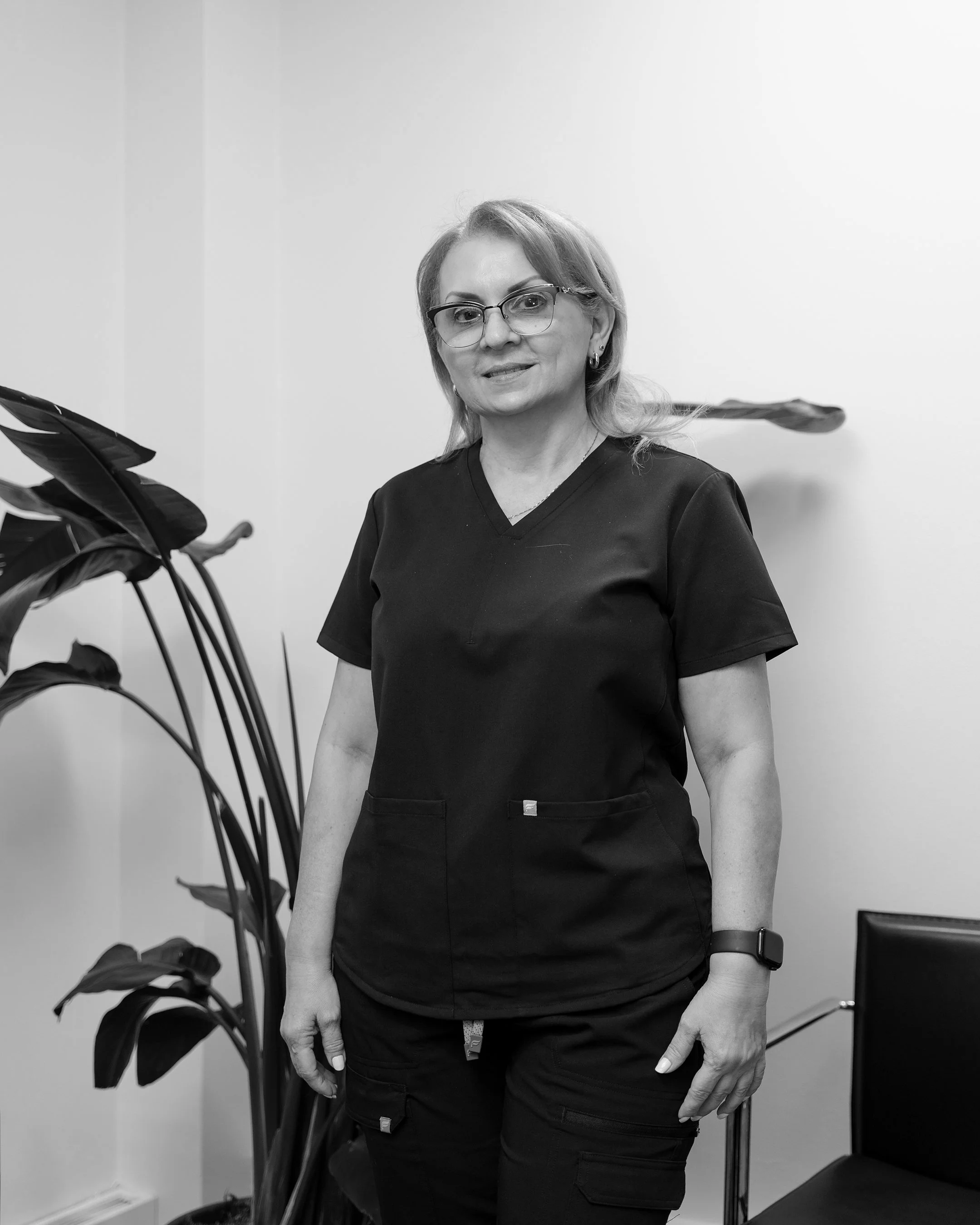Black and white photo of a woman in medical scrubs, wearing glasses, standing indoors near a potted plant and a chair.