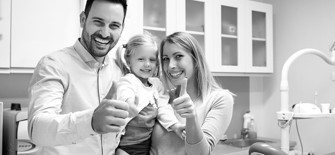 A happy family in a medical or dental office, giving thumbs up, with a man, woman, and young girl smiling at the camera.