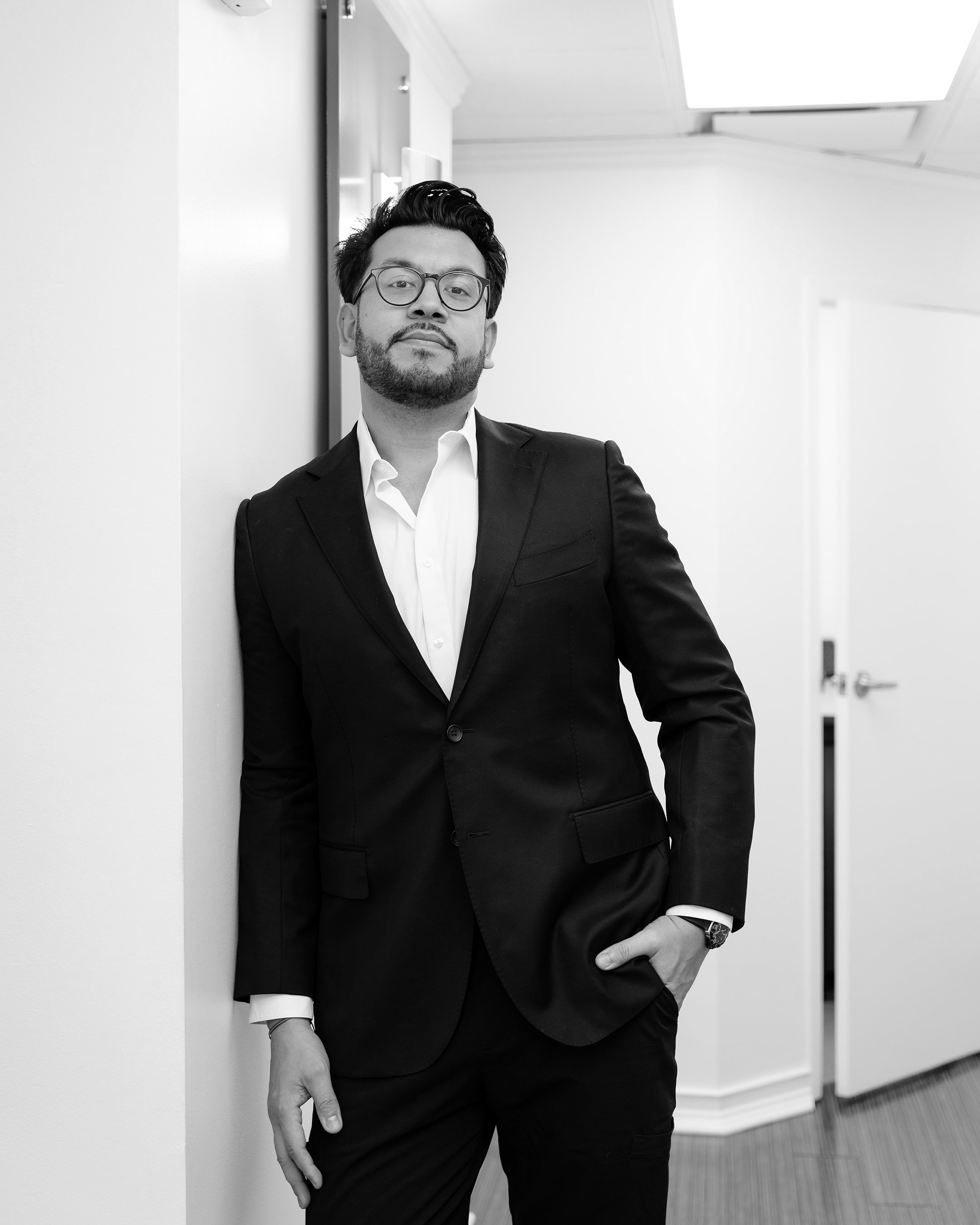 A man in a black suit and glasses leaning against a wall in a hallway, looking at the camera.
