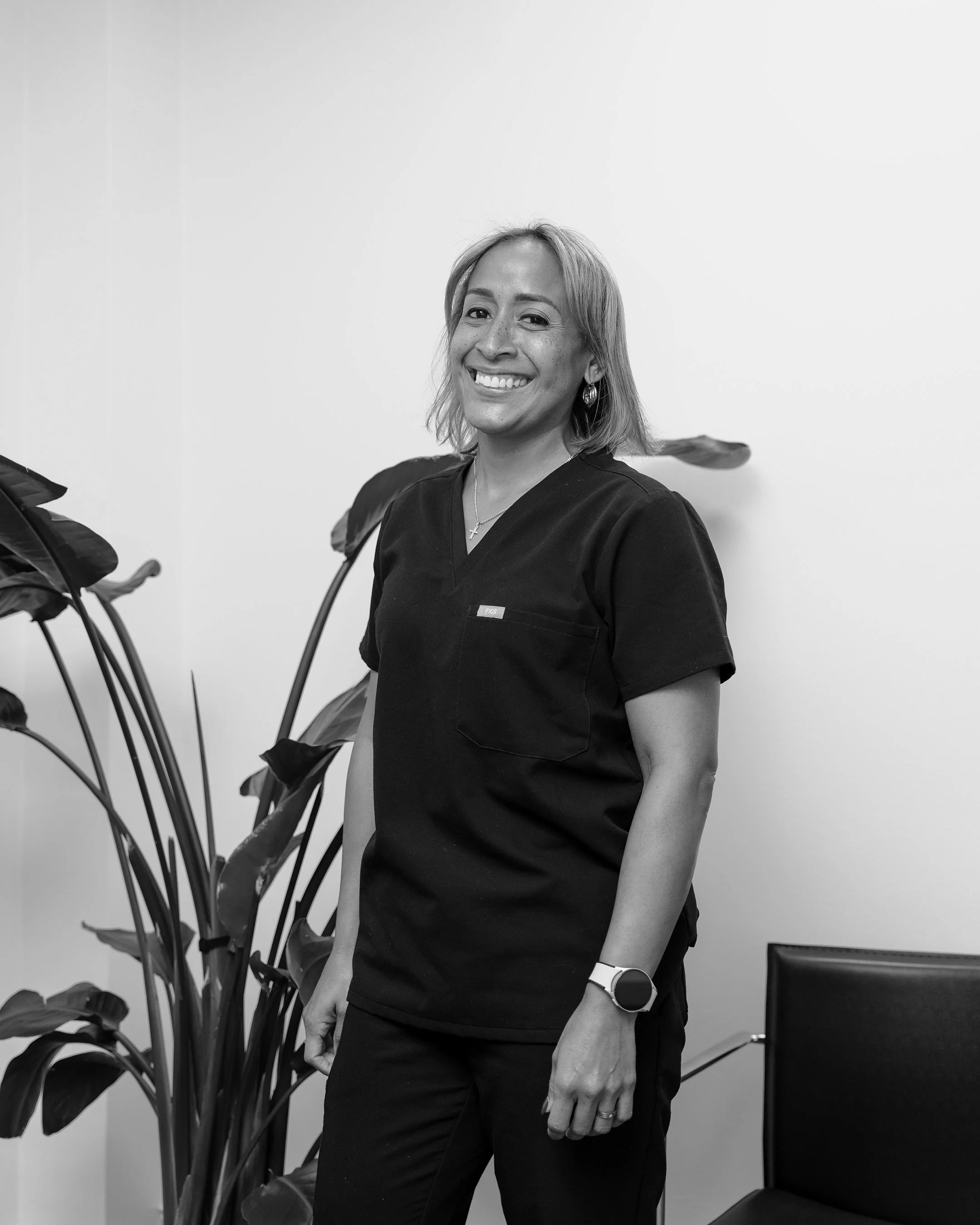 A smiling woman with shoulder-length hair wearing a black medical scrub top, standing near a large plant and a black chair against a plain white wall.