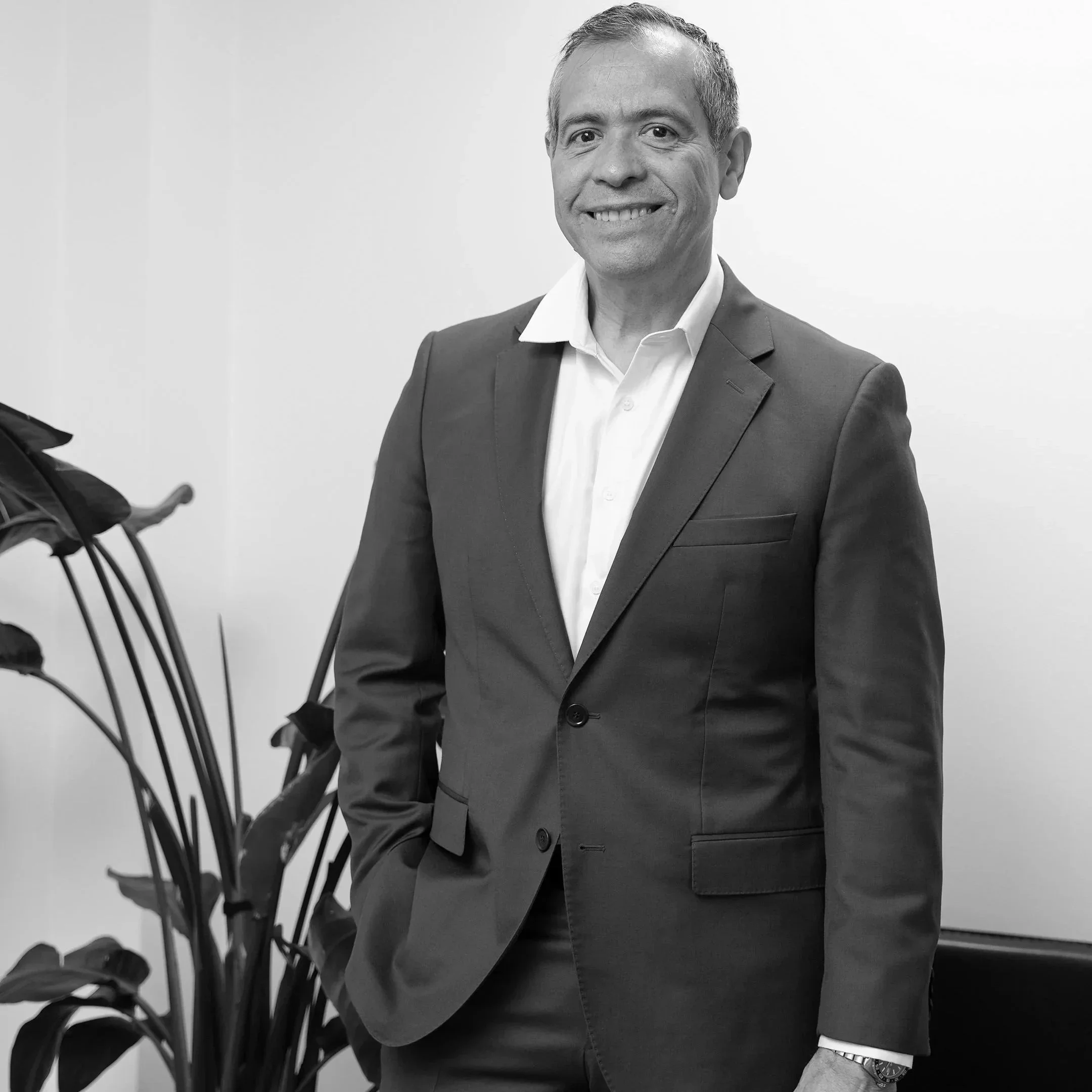 A smiling man wearing a suit and white shirt, standing next to a potted plant.