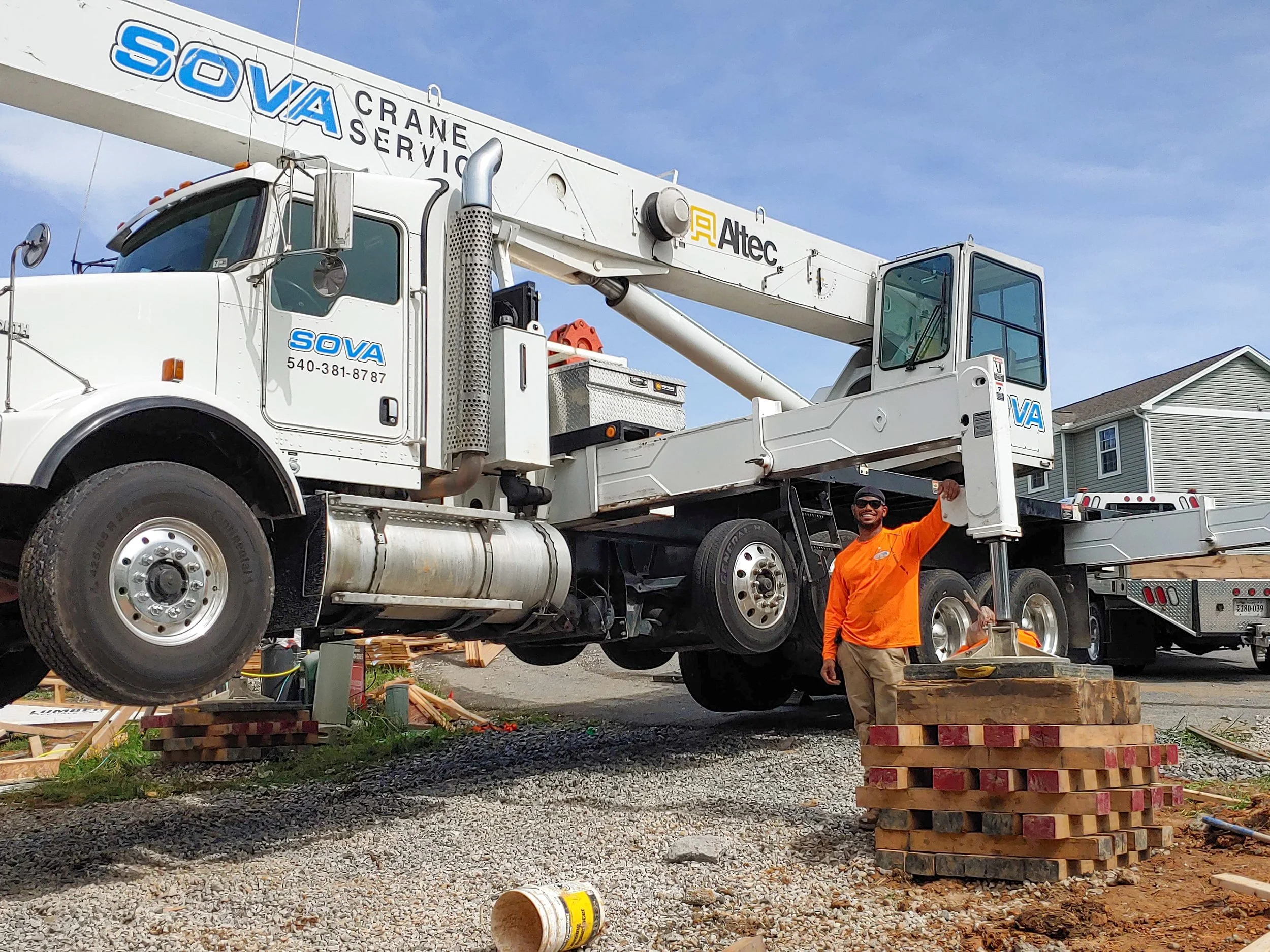 A man in orange shirt and beige pants standing next to a large white crane truck labeled SOVA, with a clear blue sky and houses in the background.