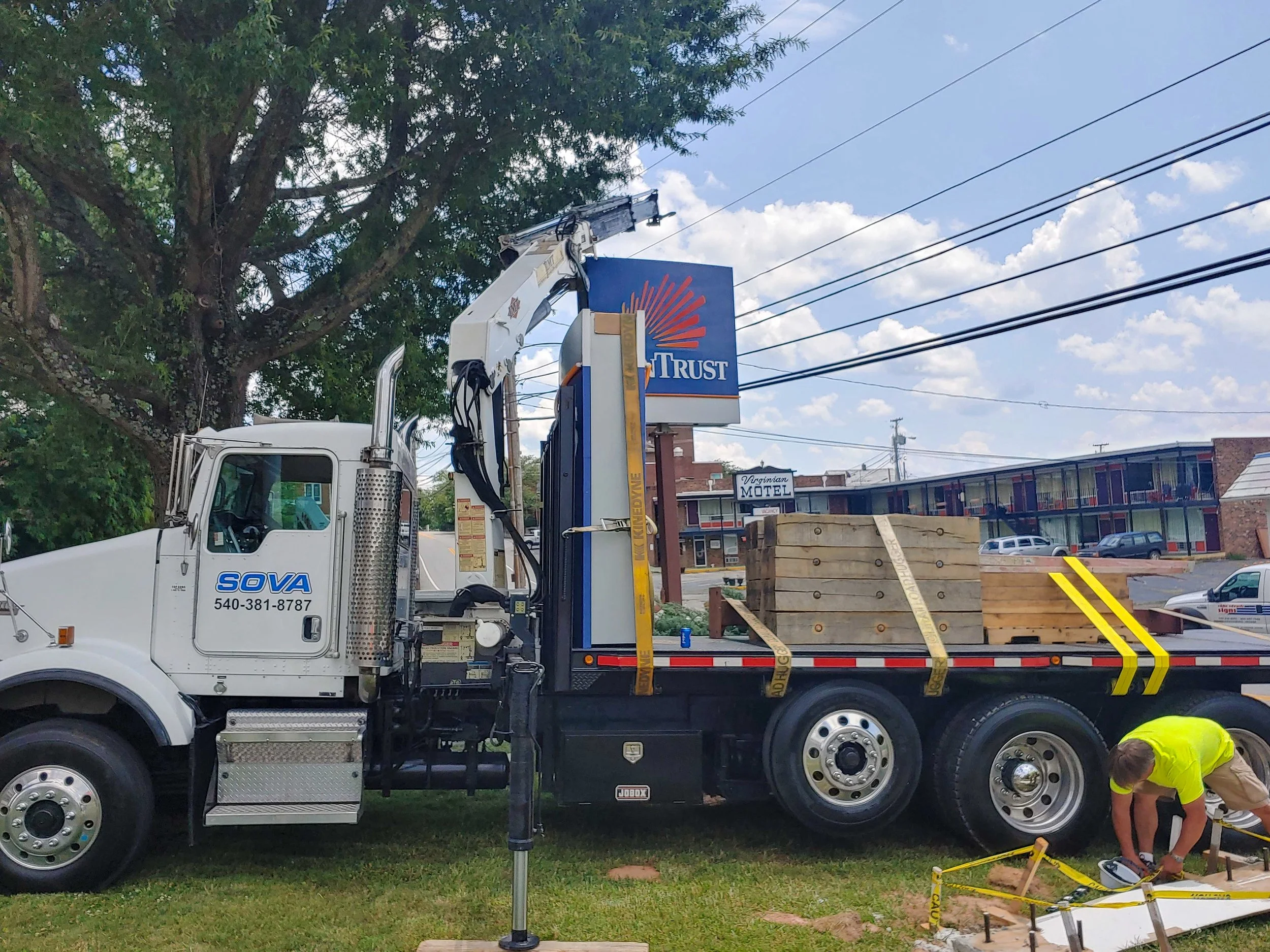 A white utility truck with a crane arm is parked on grass, with wooden crates loaded on the back. A worker in a yellow shirt is kneeling near some yellow caution tape, working on installing or inspecting equipment. In the background, trees, power lin