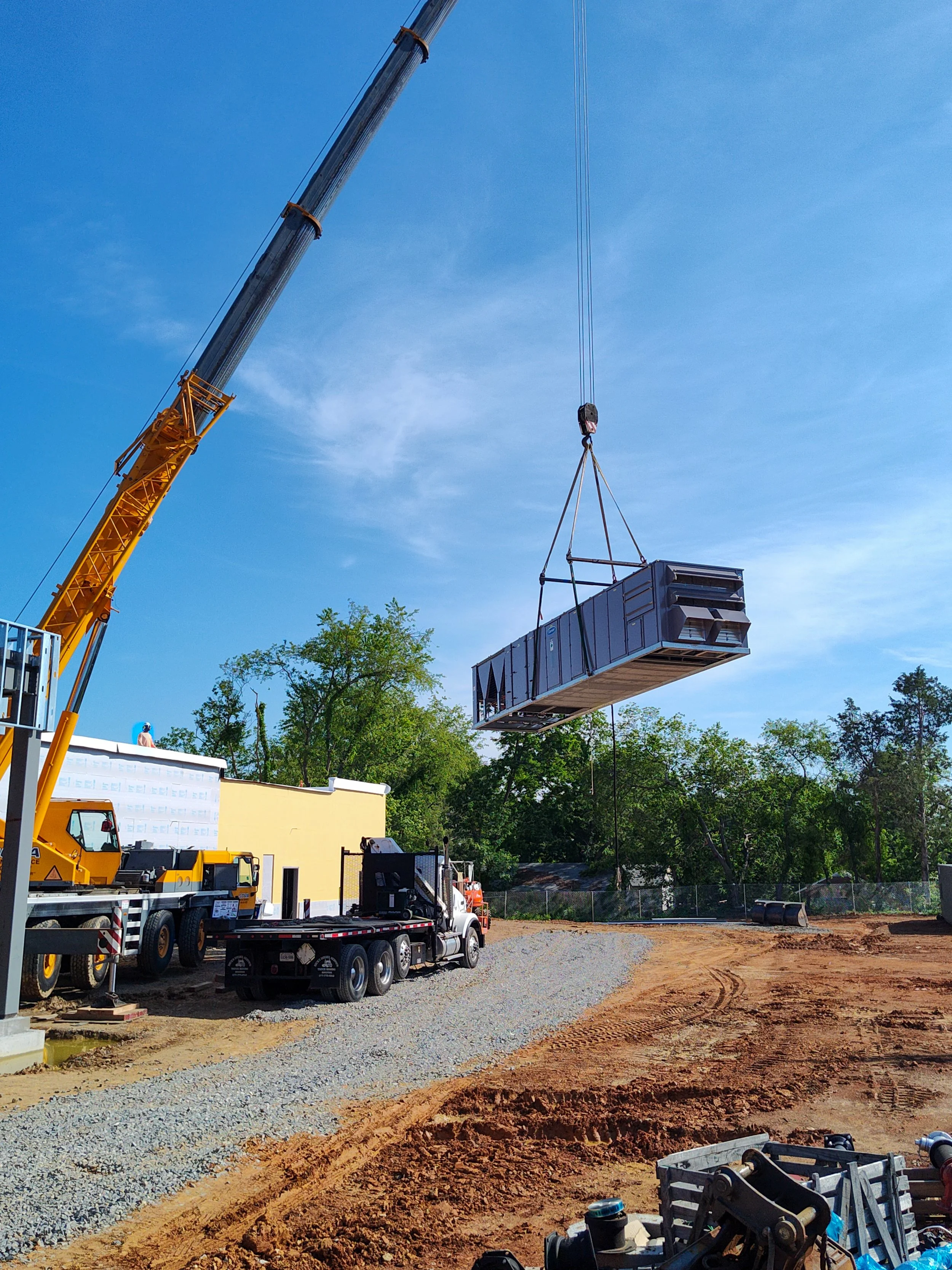 A construction crane lifting a large, dark-colored shipping container on a construction site with dirt and gravel ground, trees in the background, and a clear blue sky.