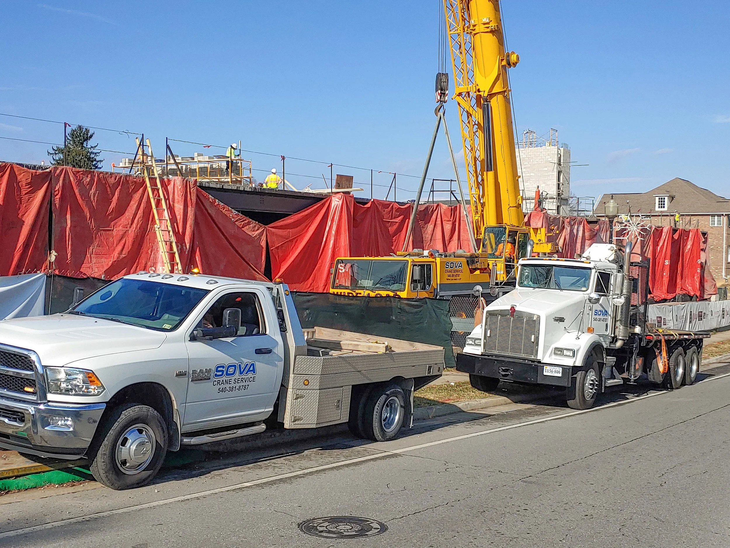 Construction site with workers on top of a building, a large yellow crane, and two trucks parked on the street in front, one with a white cab and another with a white cab and flatbed.