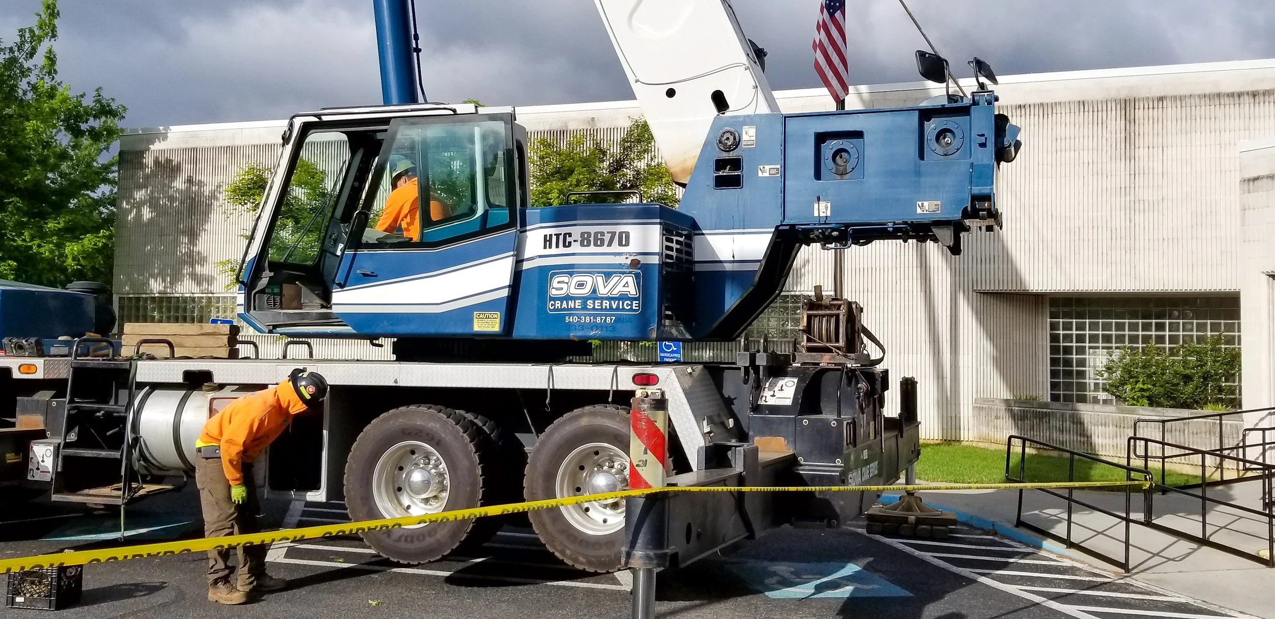 A blue crane truck with the label 'SOVA Crane Service' is parked outside a white building with glass block windows. A worker in orange shirt and safety gear is working near the truck, and caution tape surrounds the area, indicating a construction or