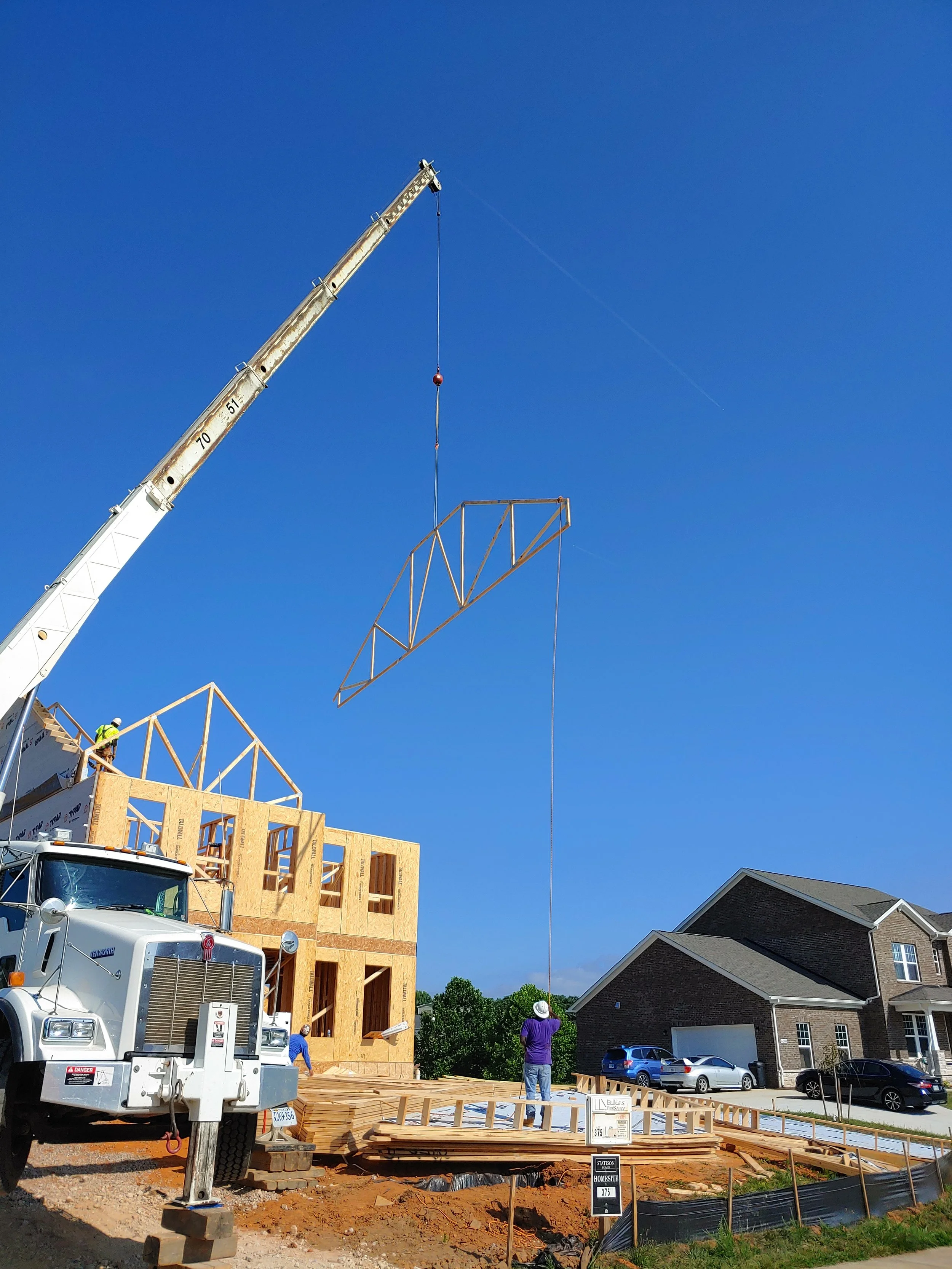 Construction site with a crane lifting building materials, partly built wooden house, and workers on site, in front of a neighboring finished house with a driveway and parked cars.