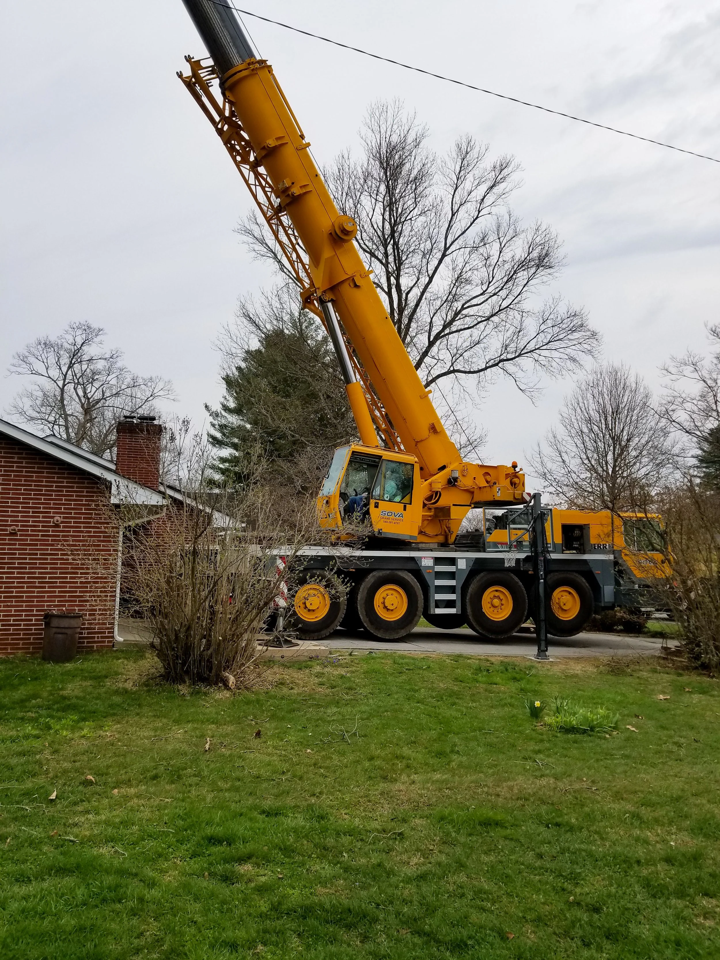 A large yellow crane parked on a driveway next to a brick house with a lawn and trees in the background.