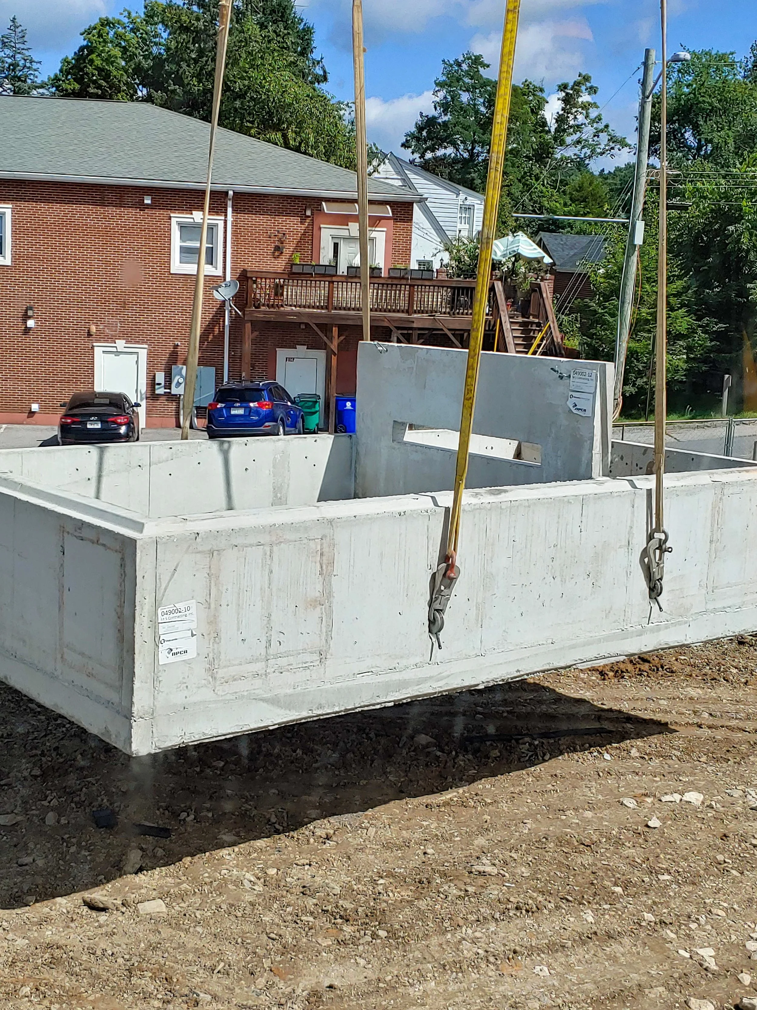 Construction site with a large concrete foundation, suspended by straps, with a residential neighborhood in the background including a brick house, parked cars, and trees.