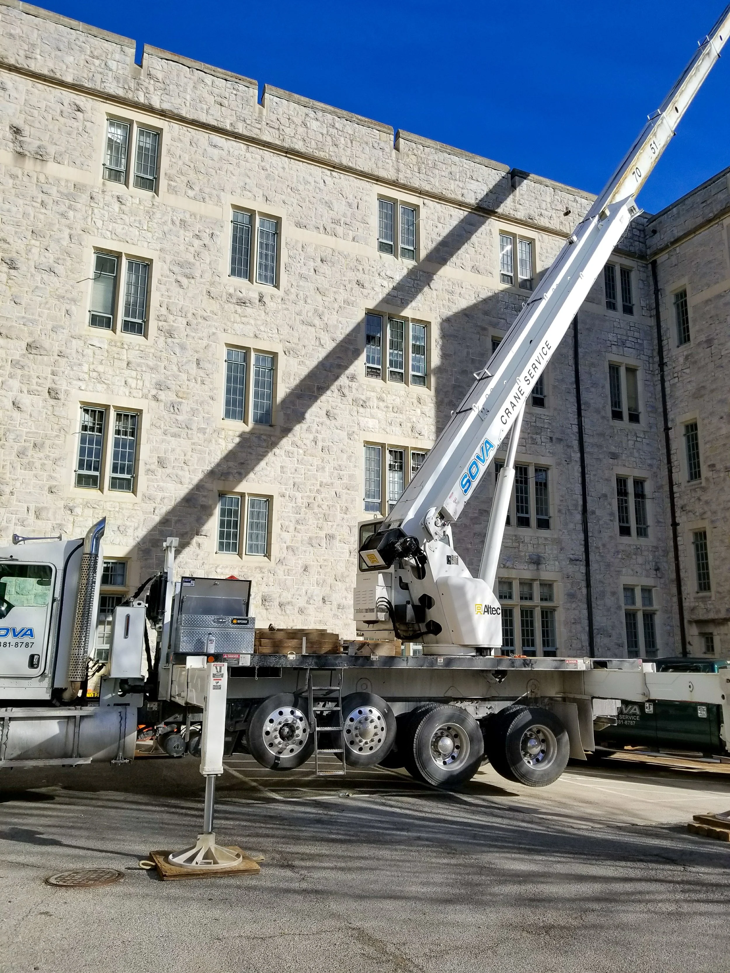 A construction crane truck is lifting or reaching above a stone building with multiple windows, with shadows cast on the wall, against a clear blue sky.