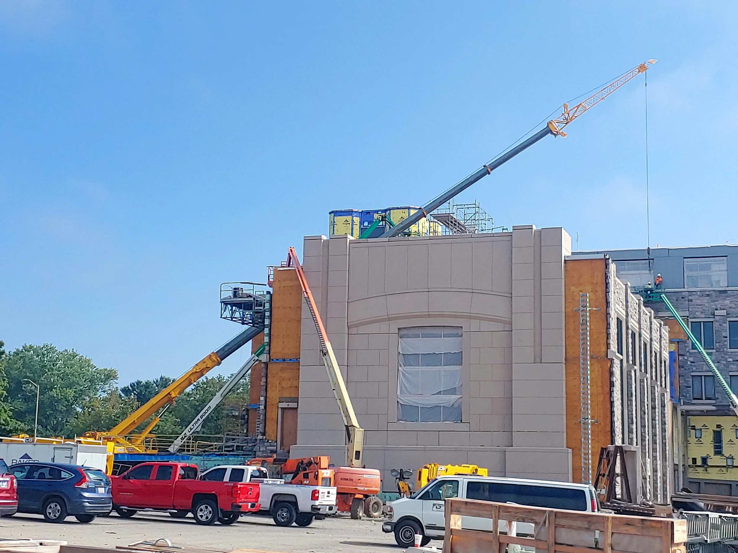 Construction site with cranes and equipment on a building under construction, parked cars in foreground, and a partly cloudy sky.
