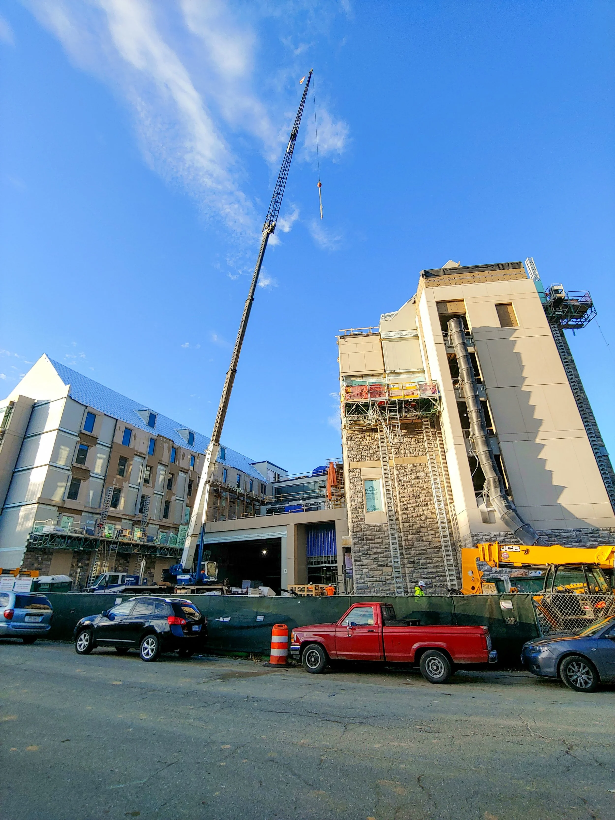 Construction site with crane and partially built multi-story building in an urban area, parked cars in the foreground, blue sky overhead.