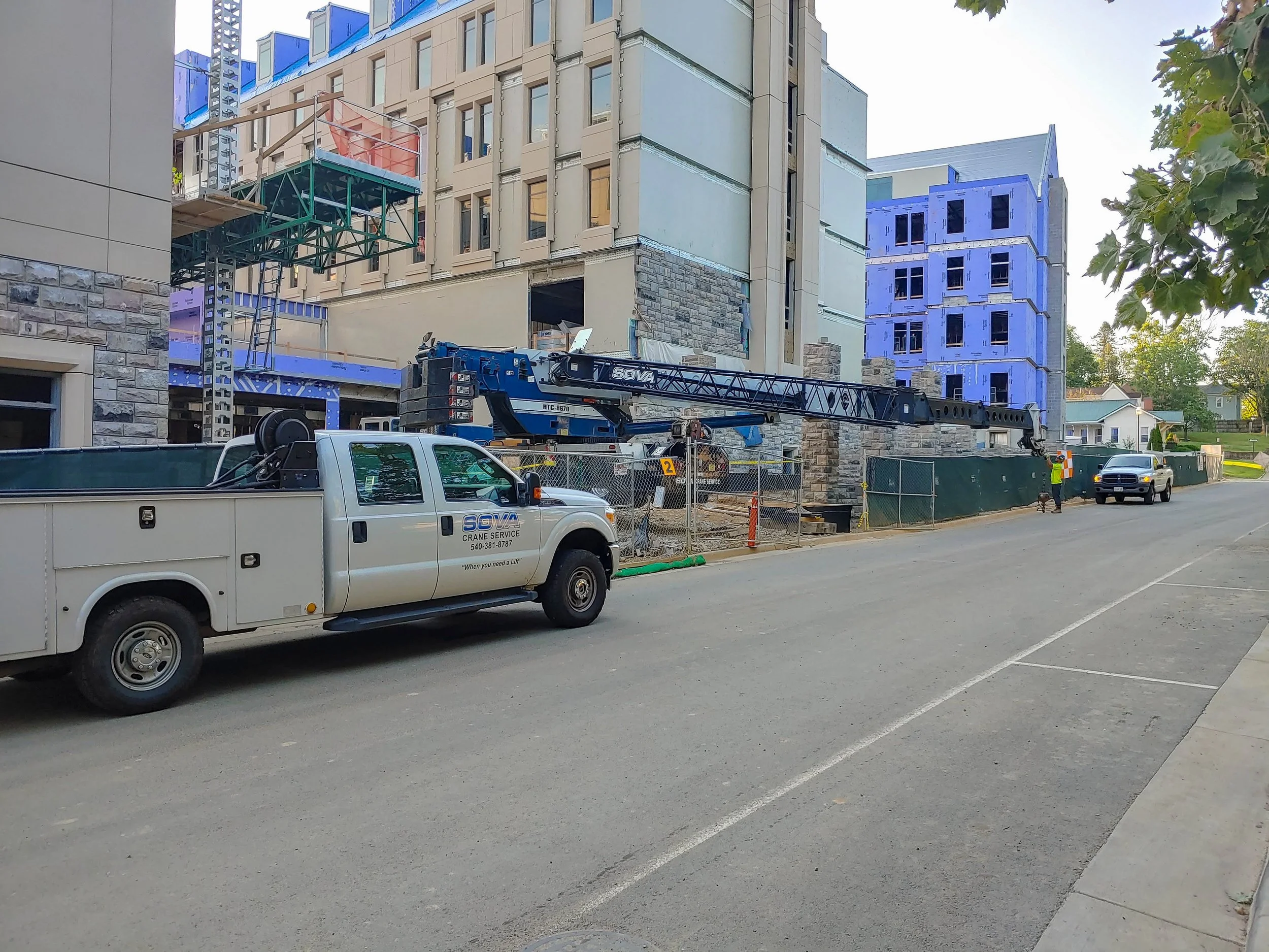 Construction site with a white crane truck parked on the road, building materials, and workers present, with a multi-story building under construction in the background.