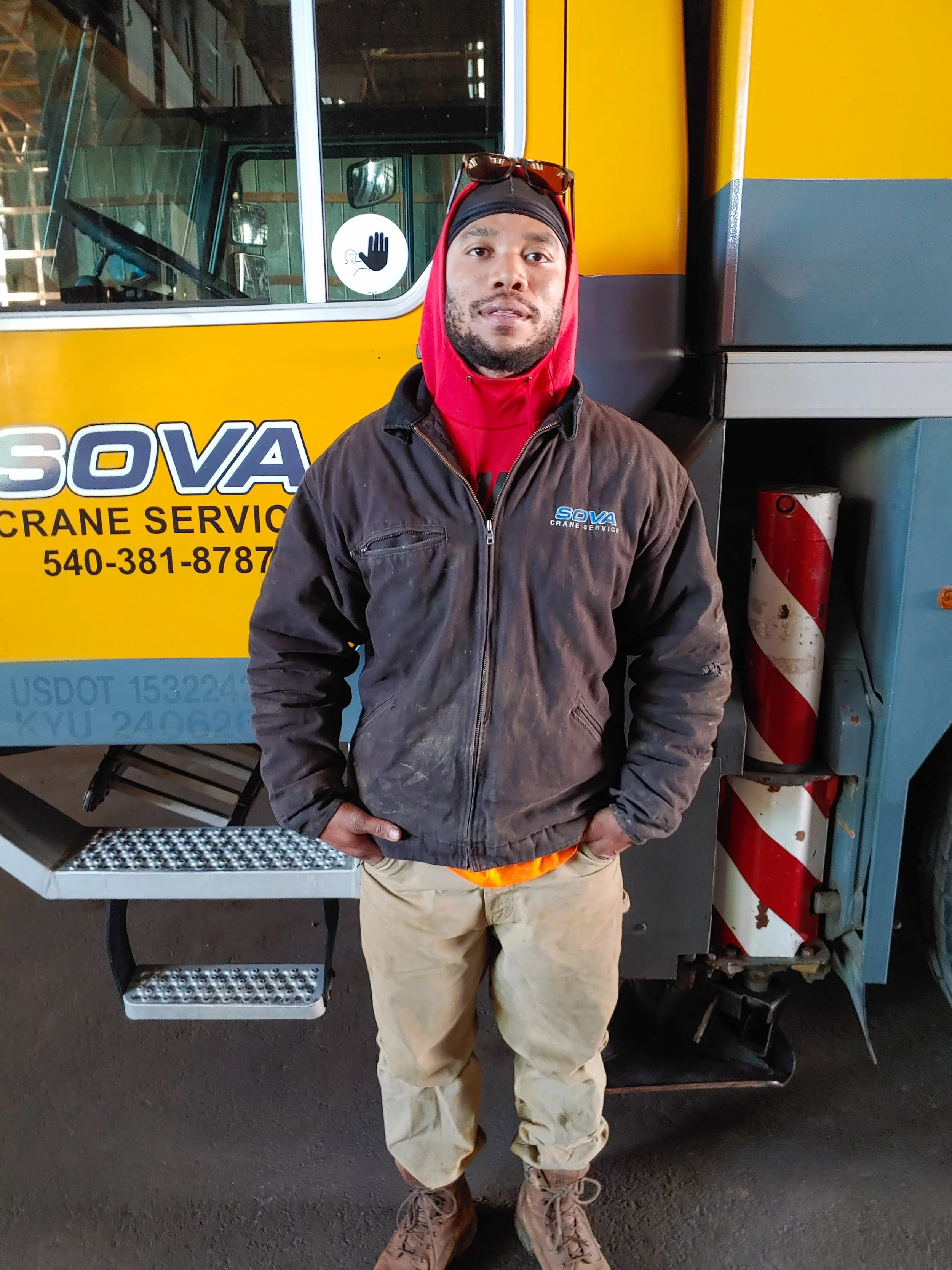 A man standing in front of a yellow crane service truck, wearing work clothes, a hoodie, and sunglasses on his head.