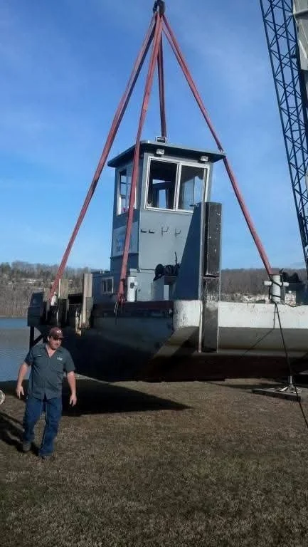 A large boat is being lifted by a crane with red straps, with a worker standing on the ground nearby wearing a dark shirt and sunglasses, outdoors on a grassy area.