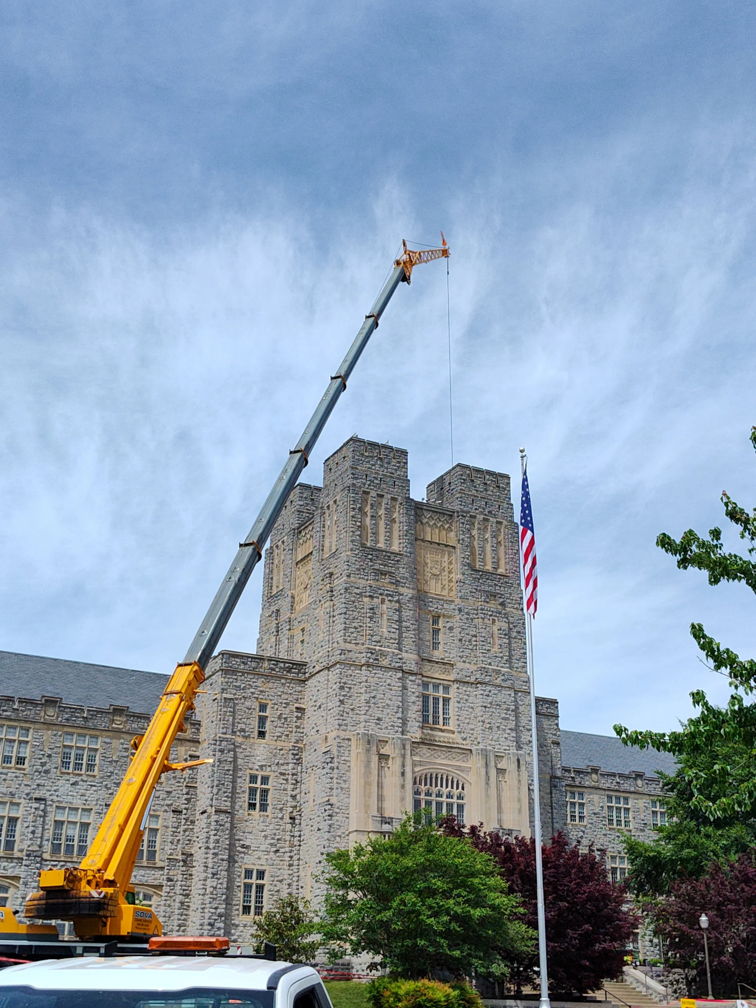 A crane working on a historic stone building, with an American flag on a flagpole and trees in the foreground.