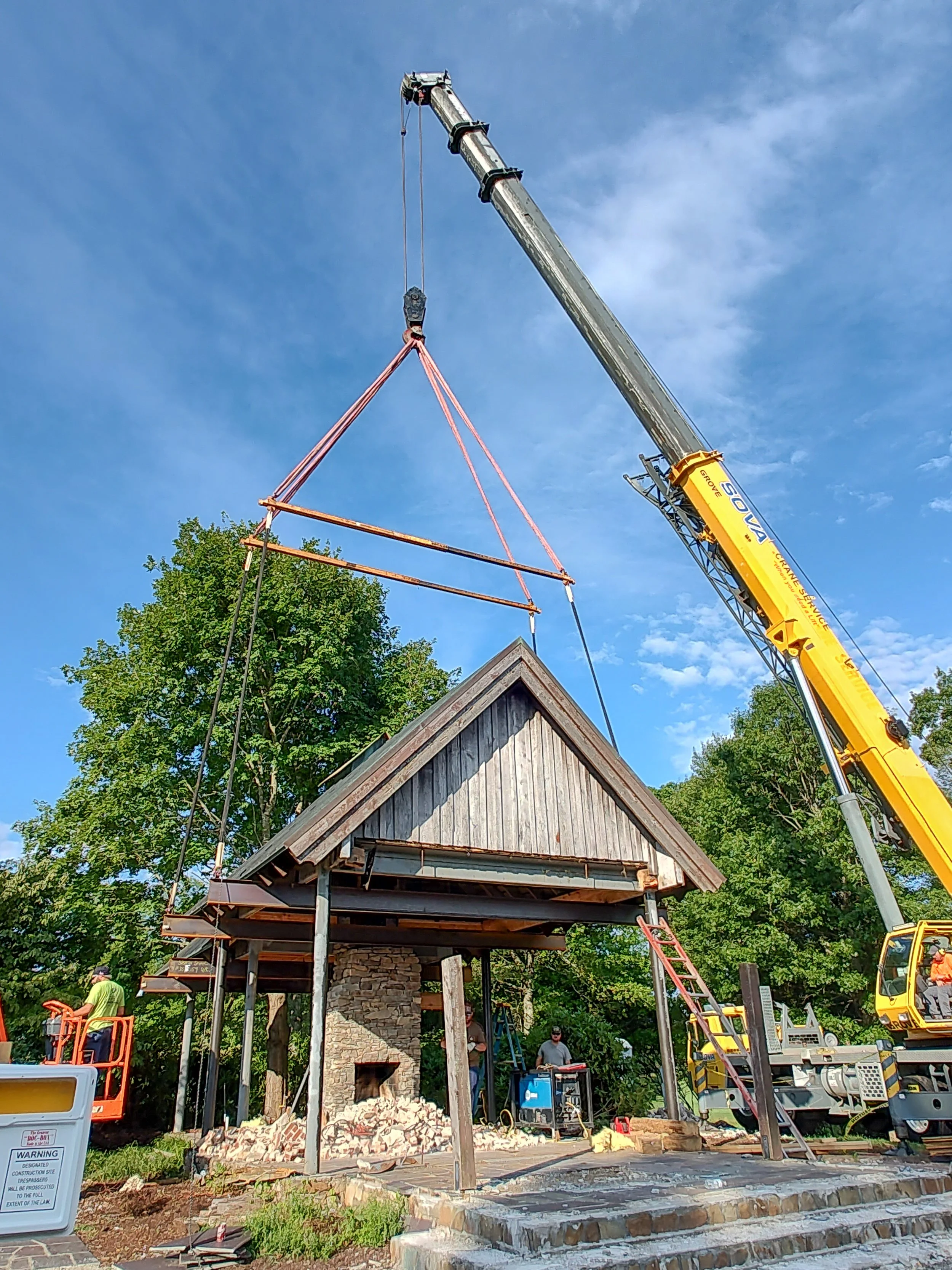 Construction workers installing a roof on a small structure with a stone fireplace, using a crane to position roofing materials, surrounded by trees and construction equipment.