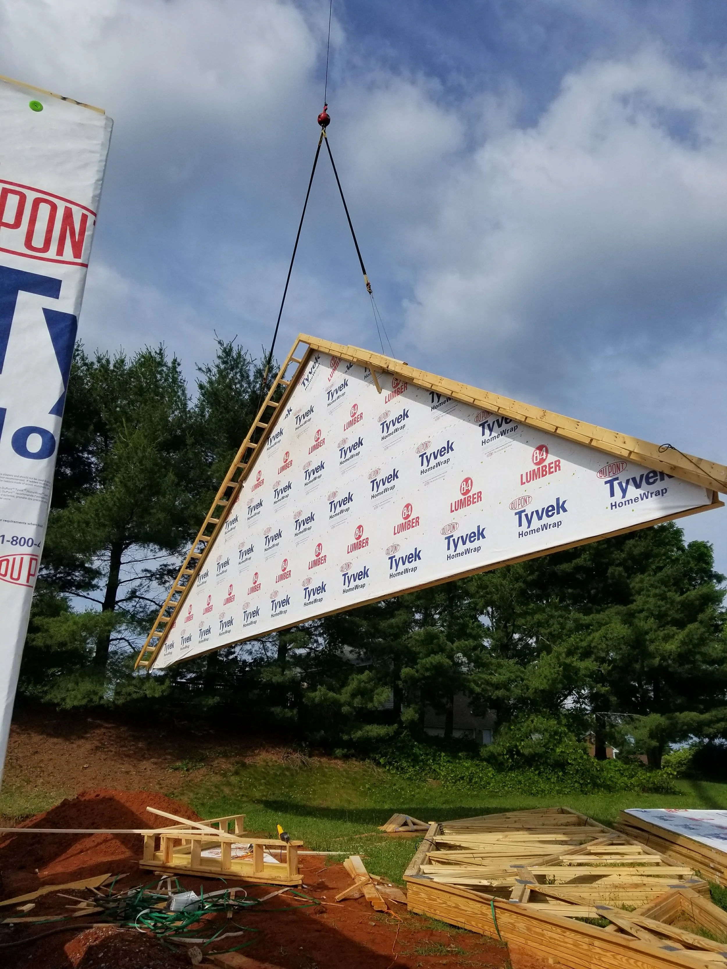 A framed wall section being hoisted into place at a construction site, with wood and dirt on the ground and some trees and a cloudy sky in the background.