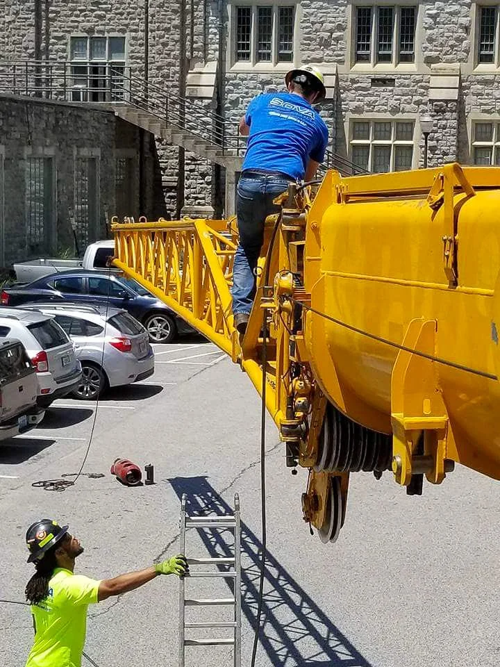 A worker in a blue shirt and helmet is climbing an extension ladder positioned on a yellow construction crane. Another worker in a neon yellow shirt and helmet is standing on the ground, holding the ladder steady. There are parked cars and an old sto