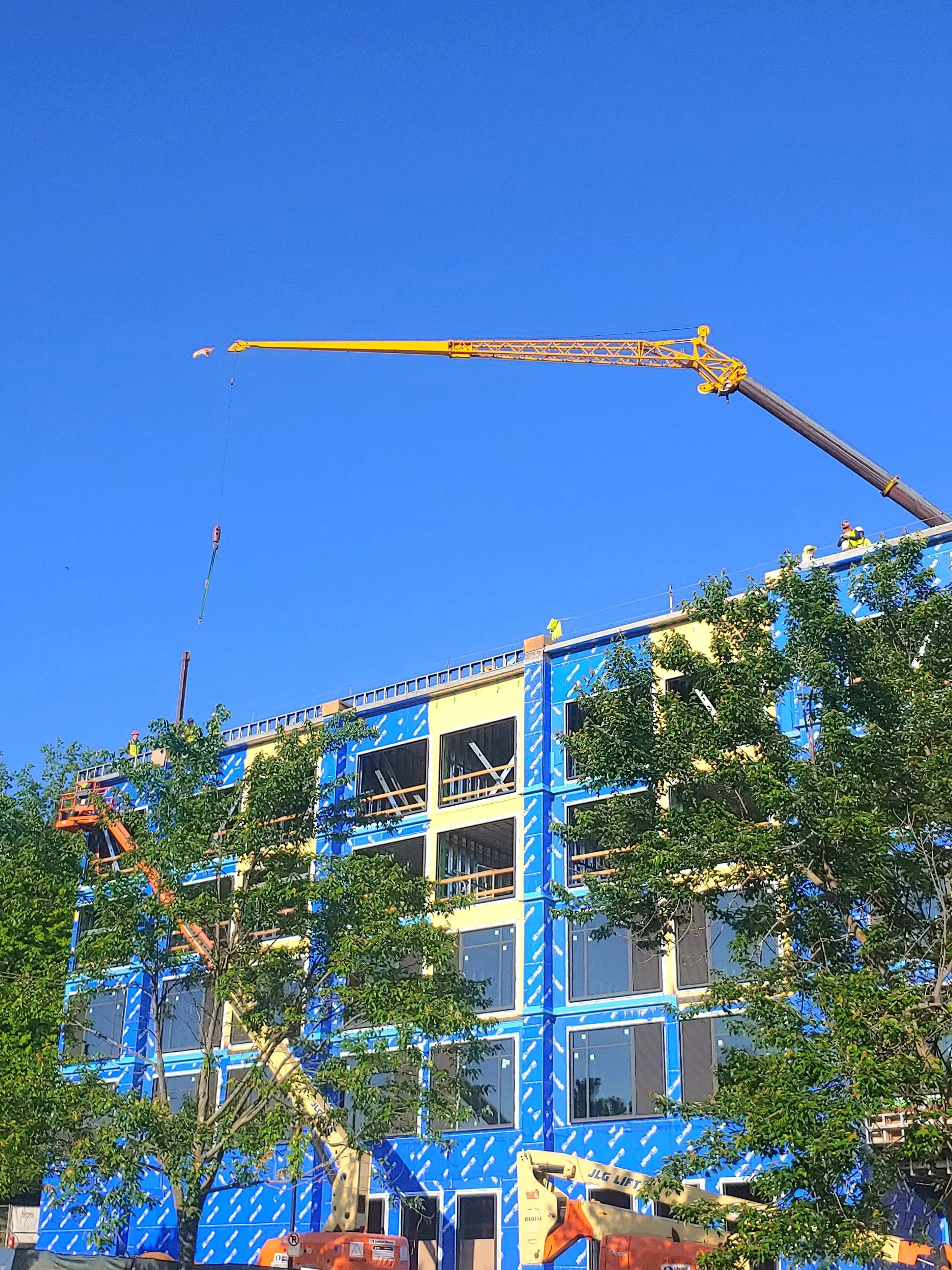 Construction site with a building in progress, surrounded by trees, featuring a tall yellow crane and construction workers on its blue exterior.