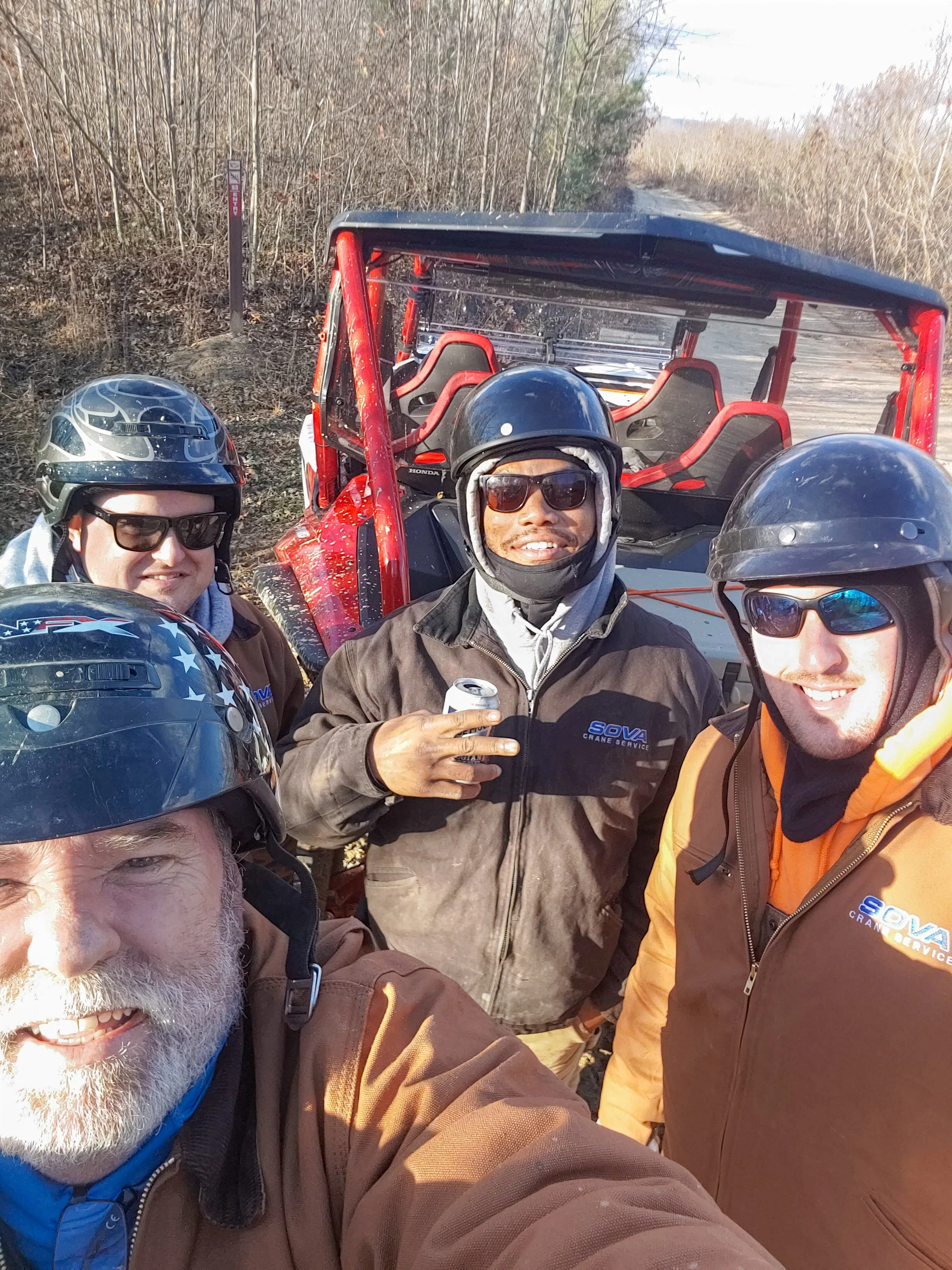 Four men wearing motorcycle helmets and sunglasses taking a selfie in front of a red all-terrain vehicle (ATV) on a wooded trail. The man in the front left has a white beard, and the others are smiling.