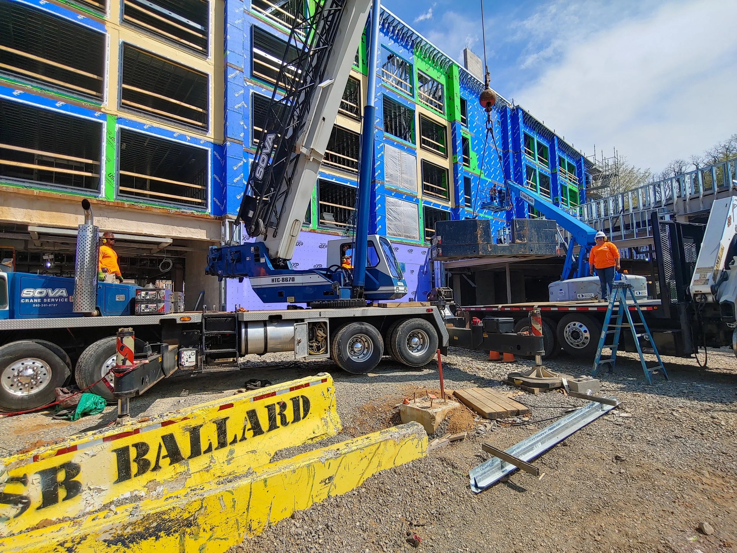 Construction workers are building a multi-story structure with blue and green exterior panels. A crane is lifting materials, and there is a truck with a lift platform. Safety barricades and construction tools are visible on the ground.