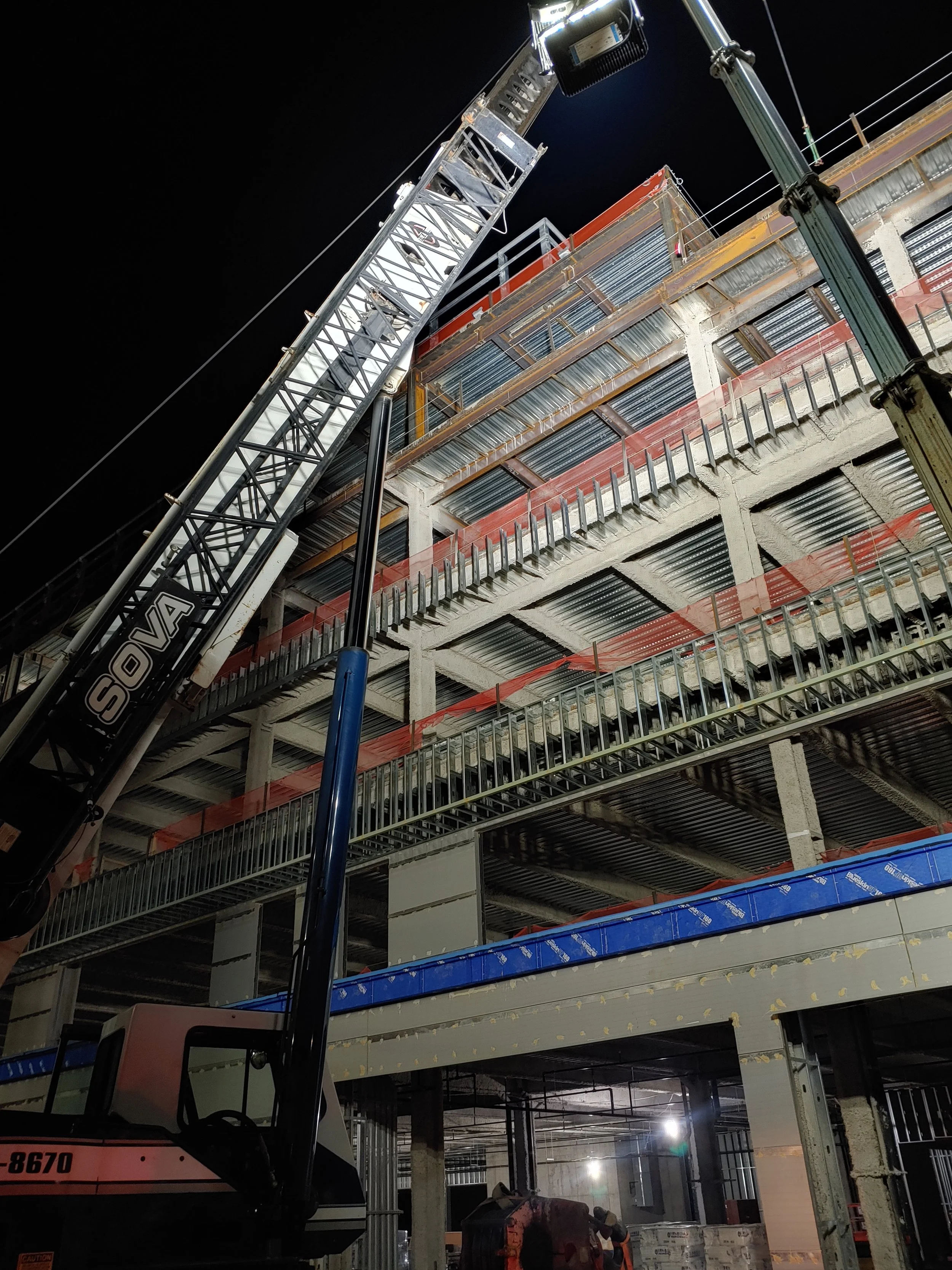Construction site at night with a crane labeled SOVA lifting materials to the upper floors of a building under construction.