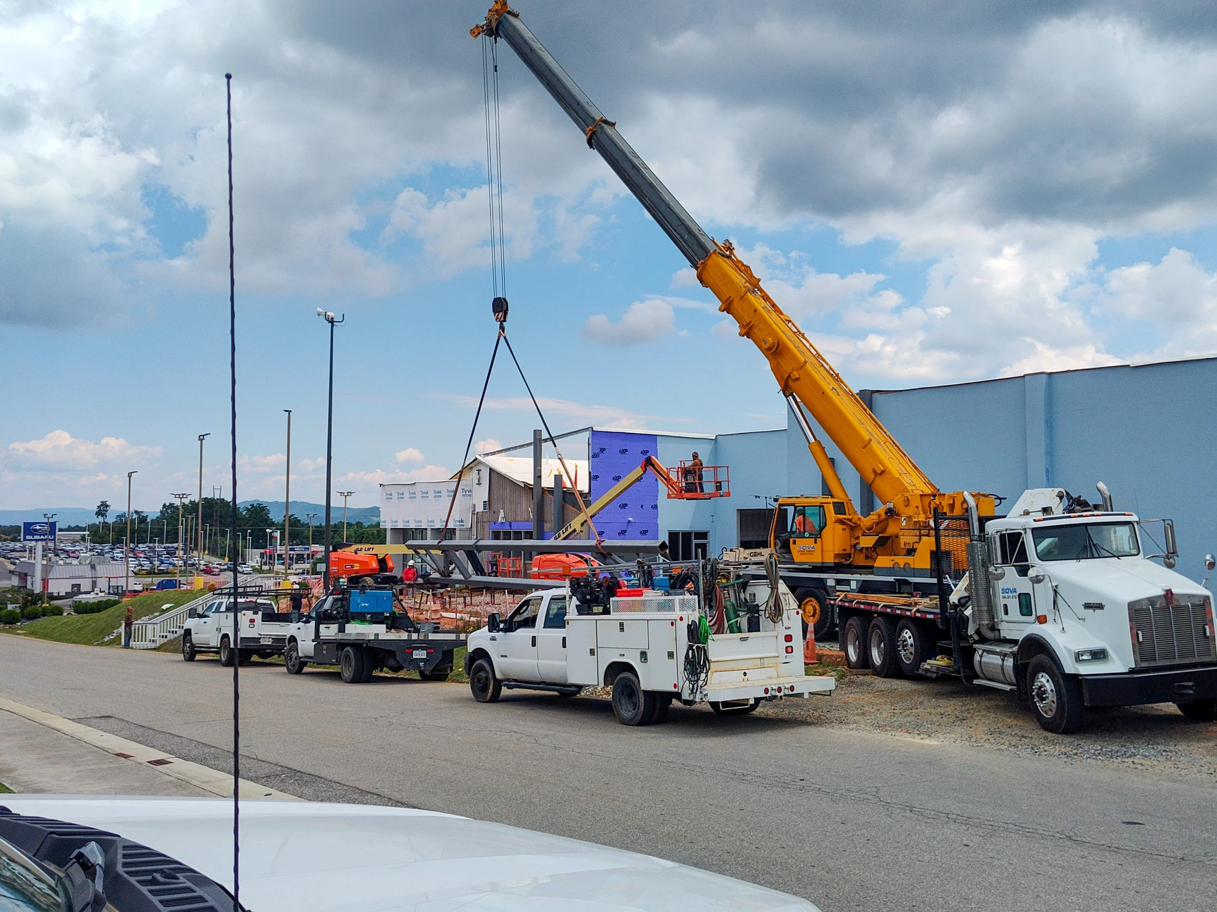 A construction site with a large yellow crane lifting a metal structure, with workmen in an elevated basket, several utility trucks parked nearby, and a partially built building in the background.