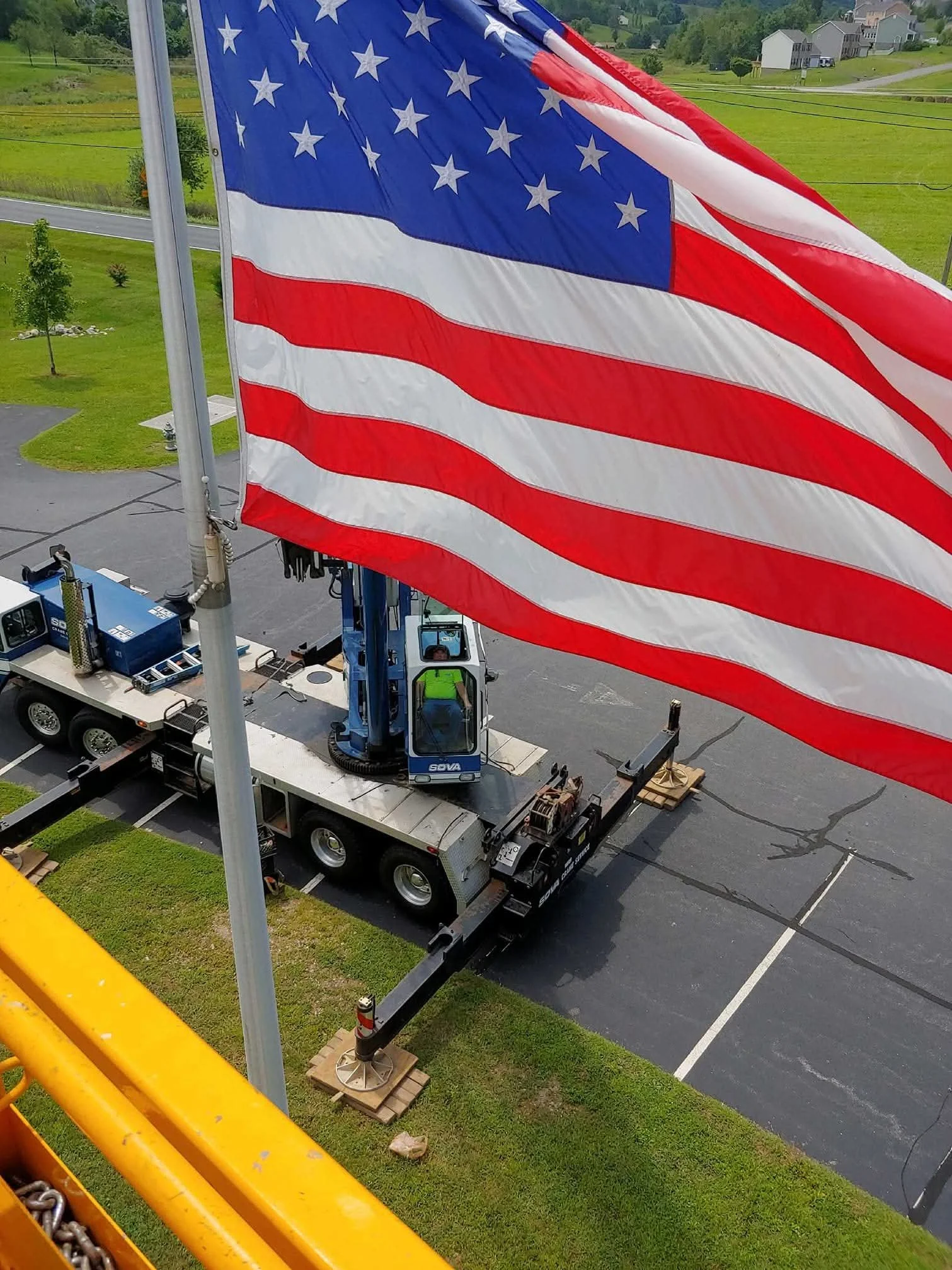Large American flag waving in the wind, mounted on a pole in a parking lot, with a construction crane and a worker visible below. Green fields and houses in the background.