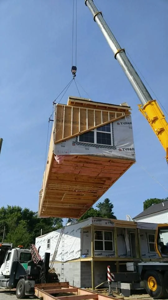 A construction crane lifting a tiny house construction section above a house on the ground on a sunny day.