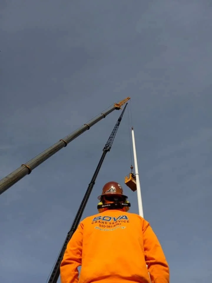 Worker in orange safety gear and hard hat overseeing a crane lifting a pole into place against a cloudy sky.