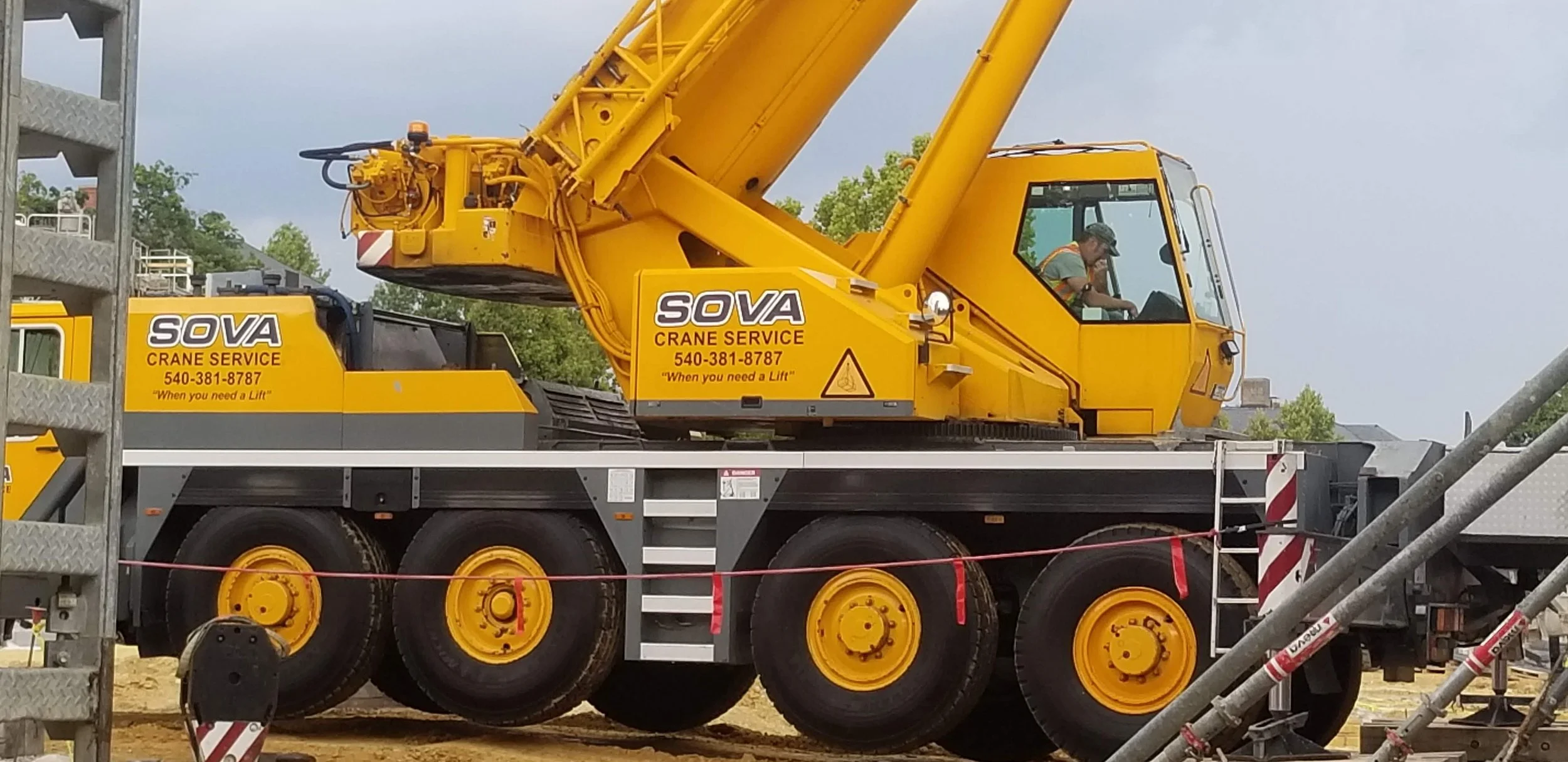 A yellow SOVA crane truck on a construction site with a worker inside the cab. The truck has multiple large black tires with yellow rims and branding on the side, including the company name SOVA, crane service contact number, and slogan 'When you nee