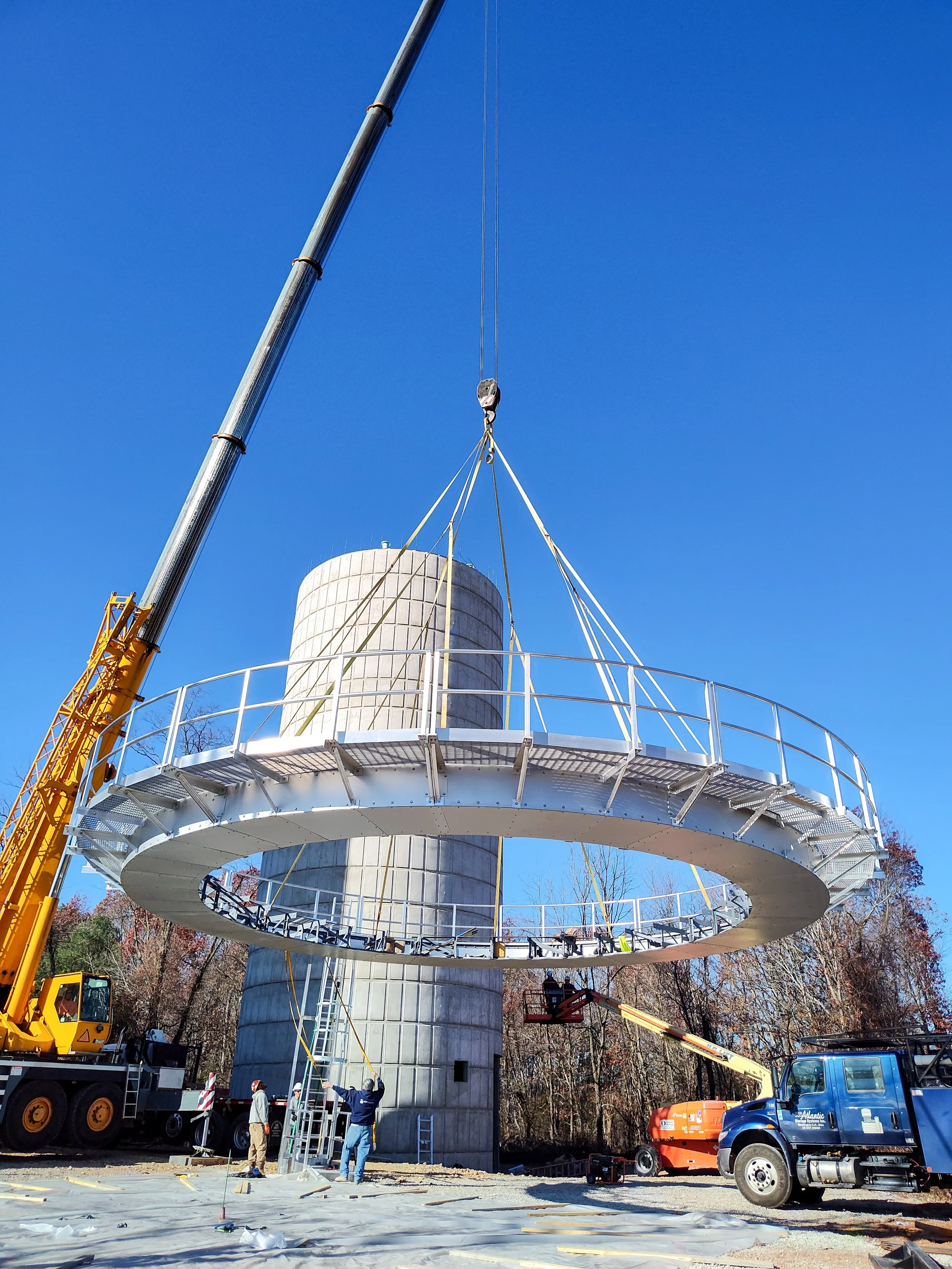 Construction workers installing a large, circular metal platform with safety railings near a cylindrical concrete tower, using cranes and lifts outdoors on a clear, sunny day.