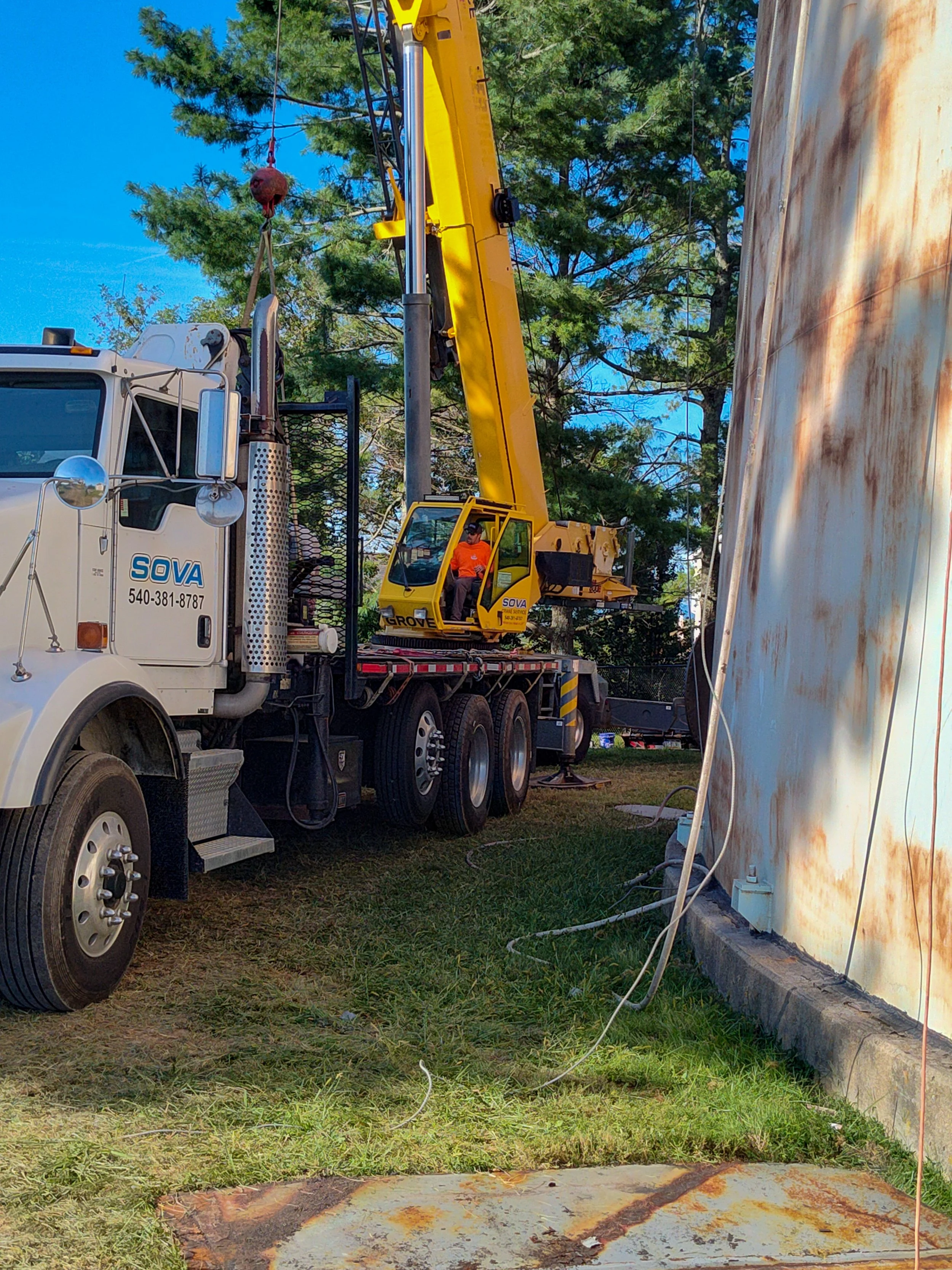 A white truck with a yellow crane attached, lifting an object near a rusty metal wall with grass and trees in the background.