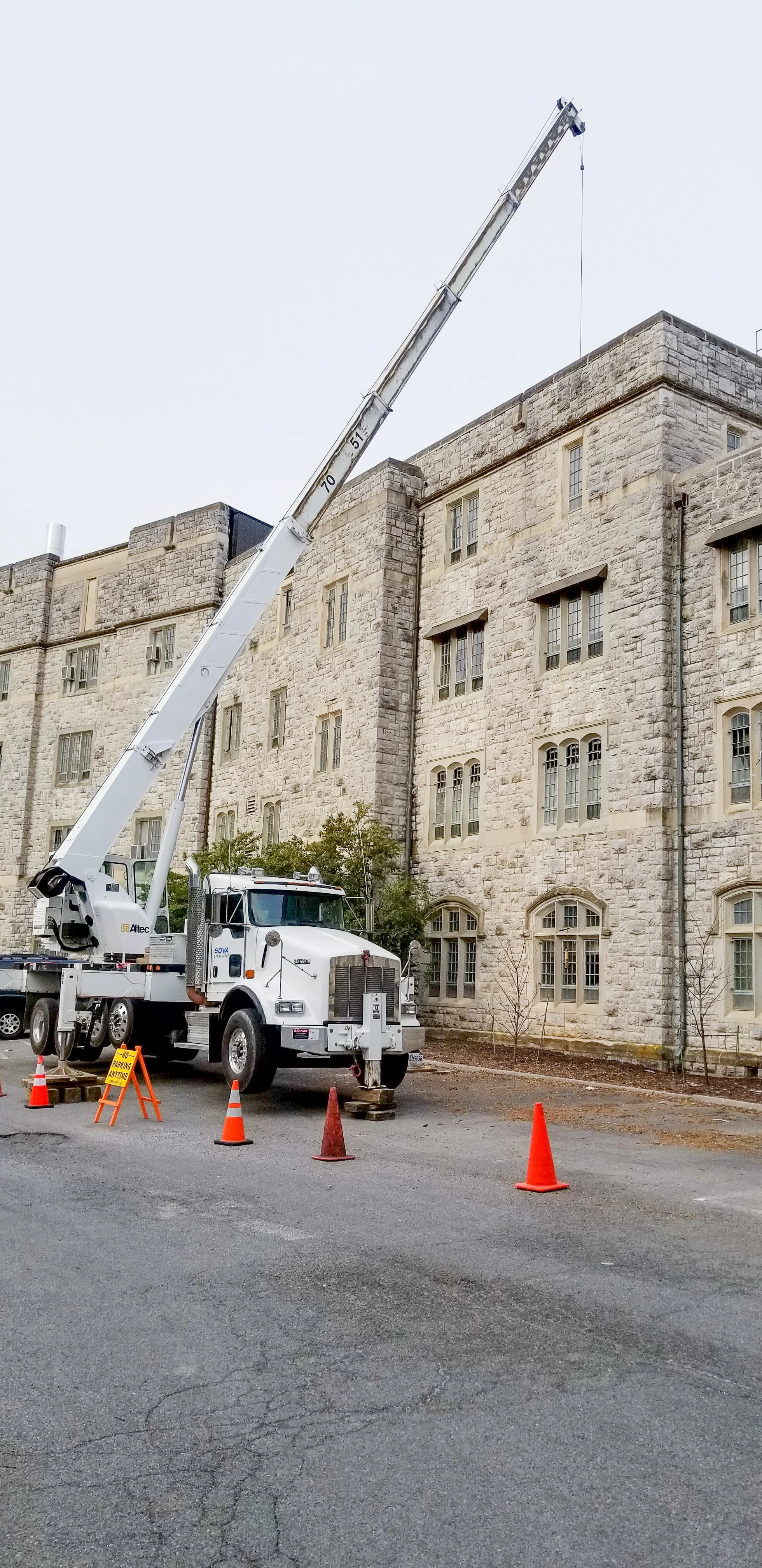A large white crane truck with an extended arm behind a historic stone building, surrounded by orange cones and a warning sign on a paved street.