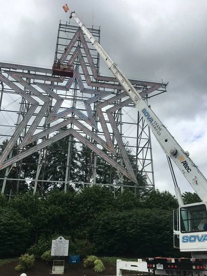 A large illuminated star structure under construction outdoors with a crane lifting part of it, against a cloudy sky and surrounded by trees and greenery.