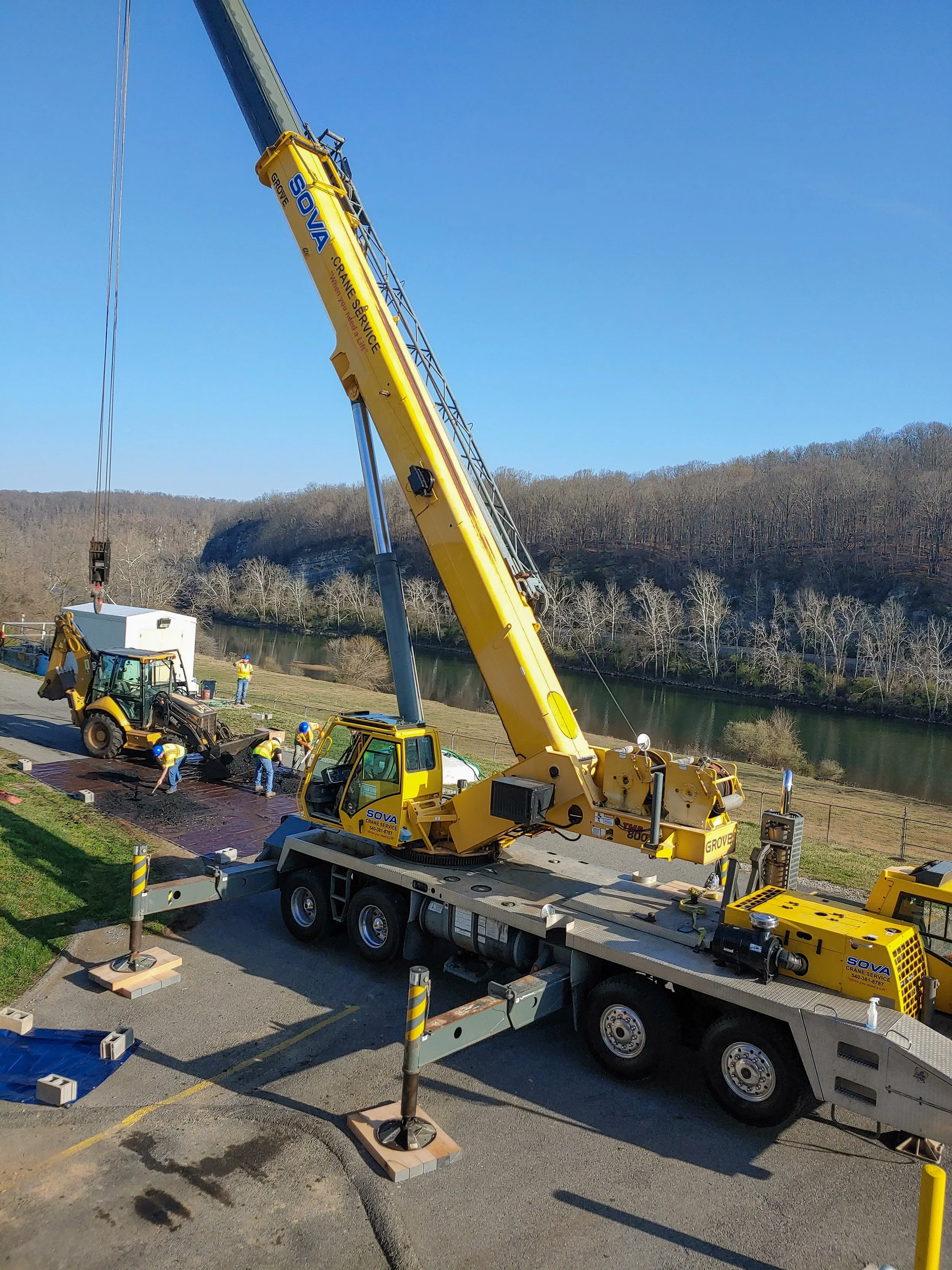 A yellow crane lifting a concrete block with workers nearby in an outdoor area by a river, with a clear blue sky and a wooded hillside in the background.