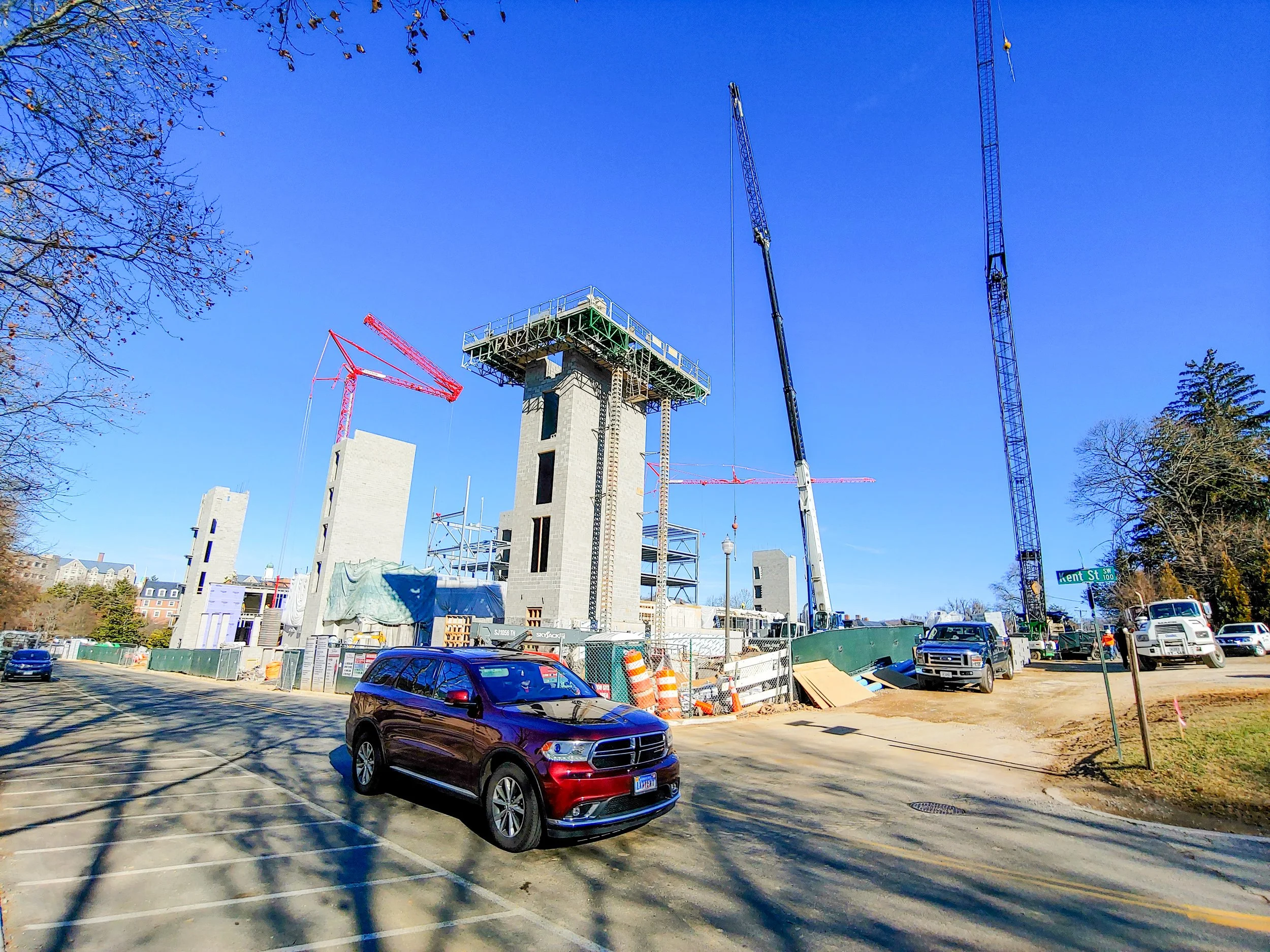 Construction site with multiple cranes and partially built concrete structures, parked cars in the foreground, and a street sign indicating 'Kent St' at the corner.