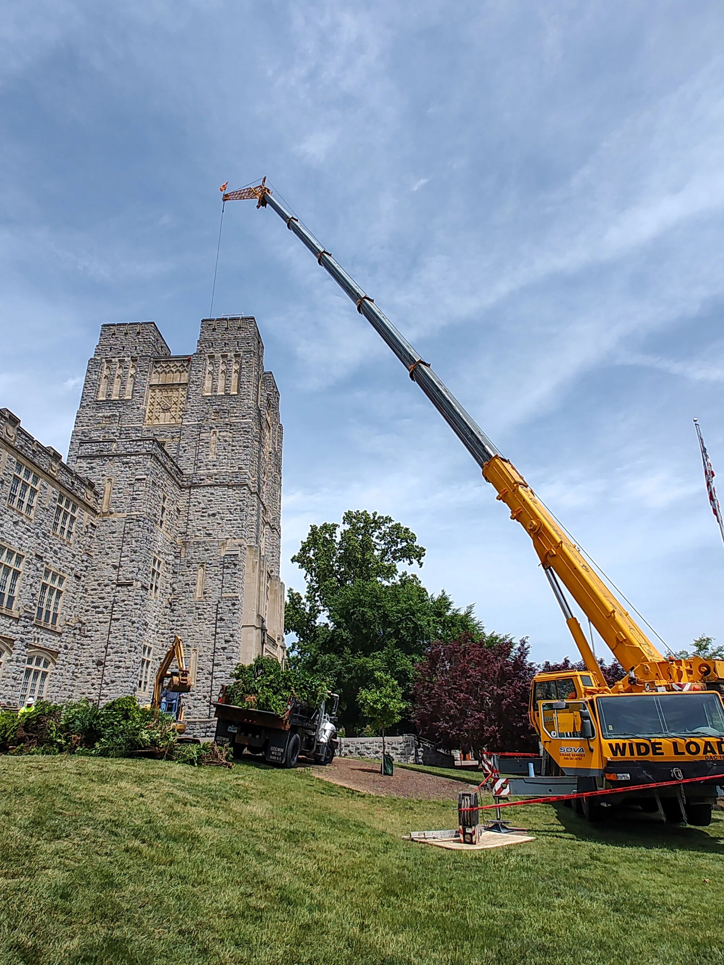 A large yellow crane with extended arm working near a historic stone building, with green grass, trees, and a blue sky with clouds in the background.