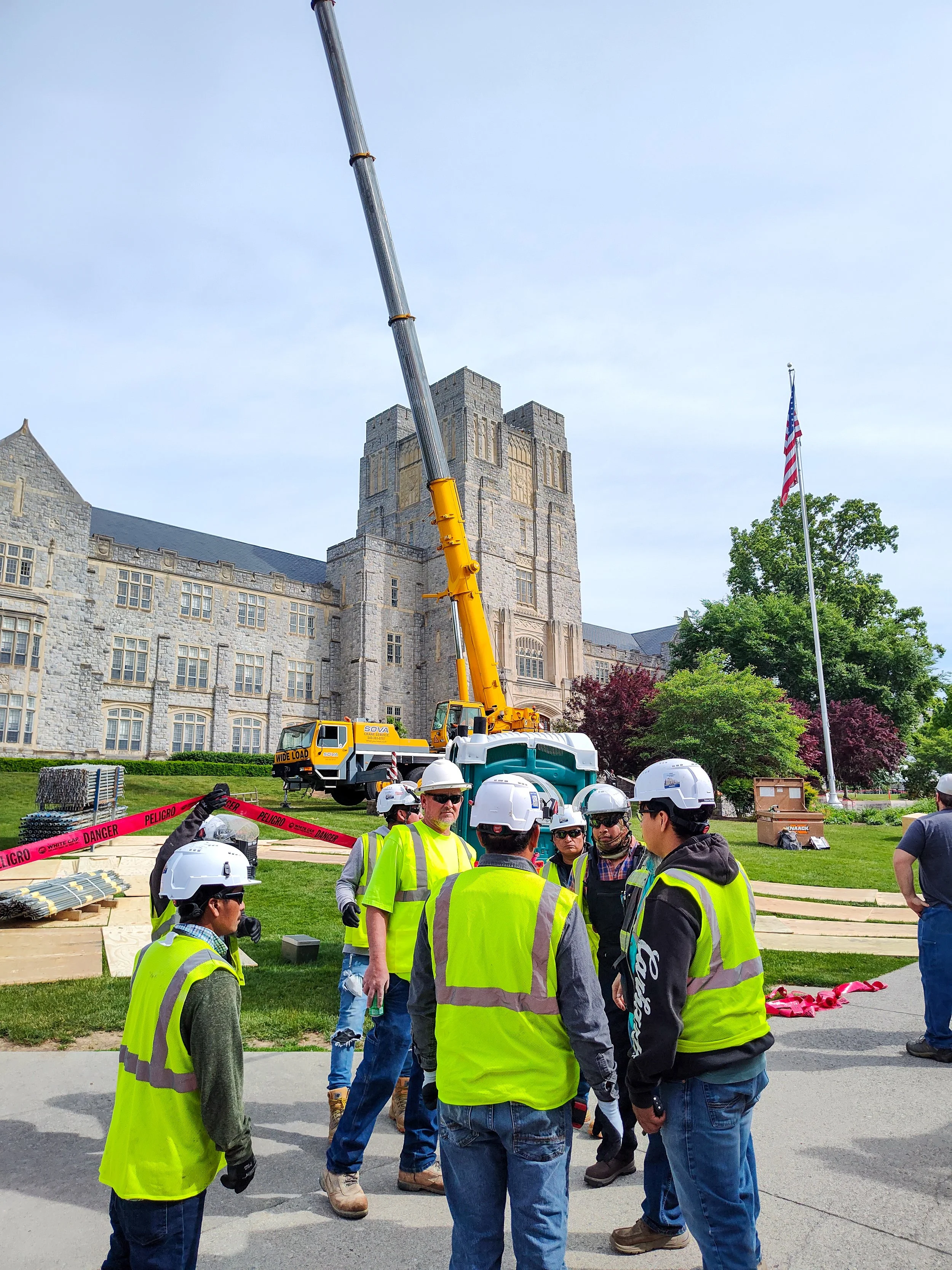 Group of construction workers with safety vests and helmets standing near a construction site in front of a historic stone building with a crane and American flag.