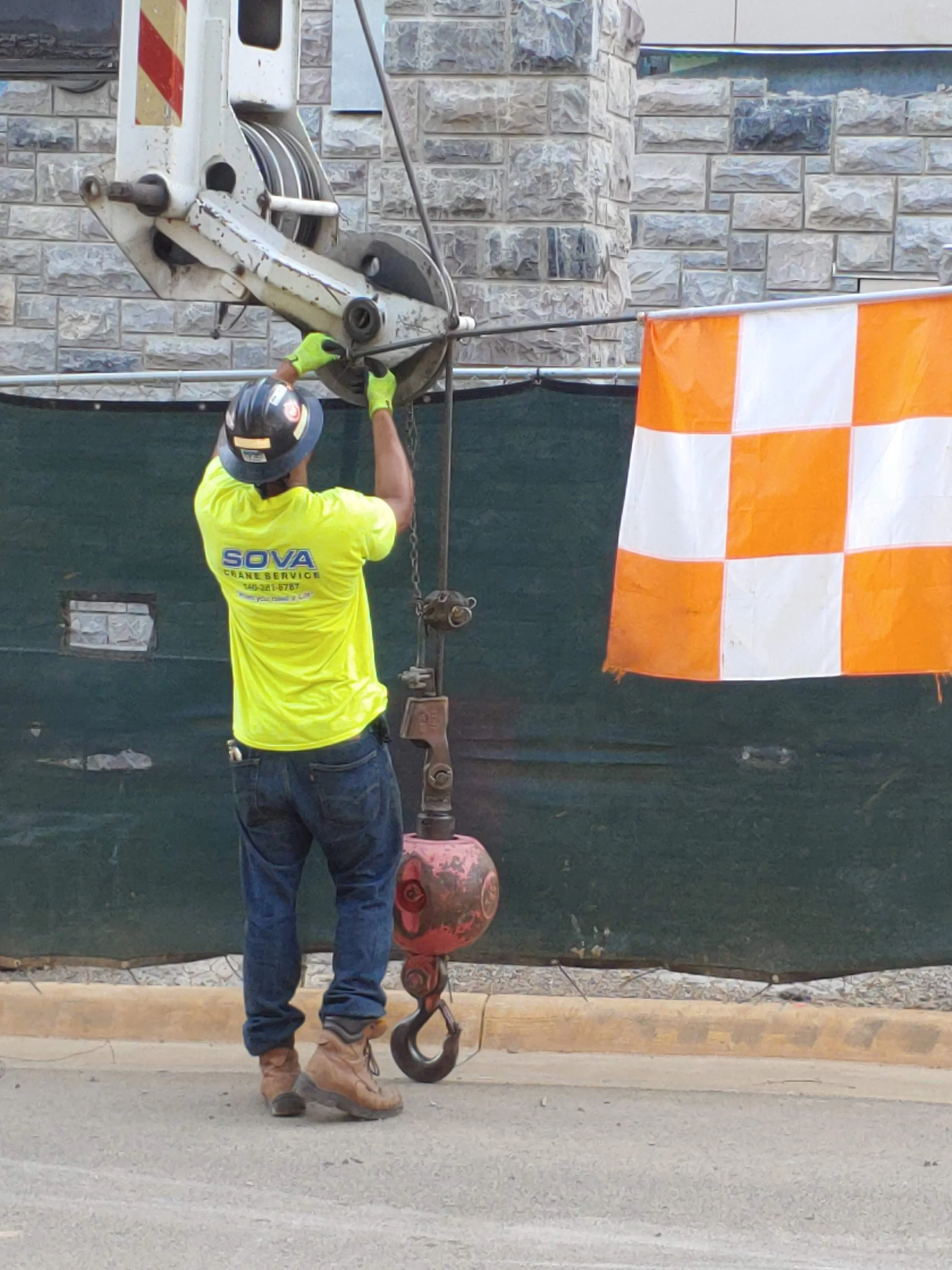 Worker in a yellow shirt and jeans operating a crane hook in front of a stone wall with an orange and white checkered flag.