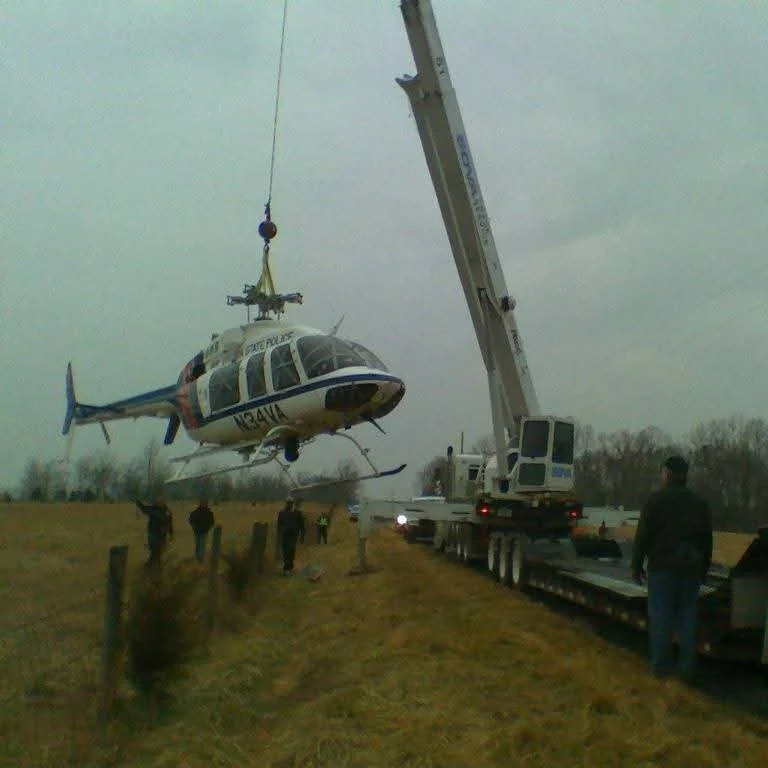 A police helicopter is being lifted or transported by a large crane on a flatbed truck, with several people standing nearby in an open field with overcast skies.