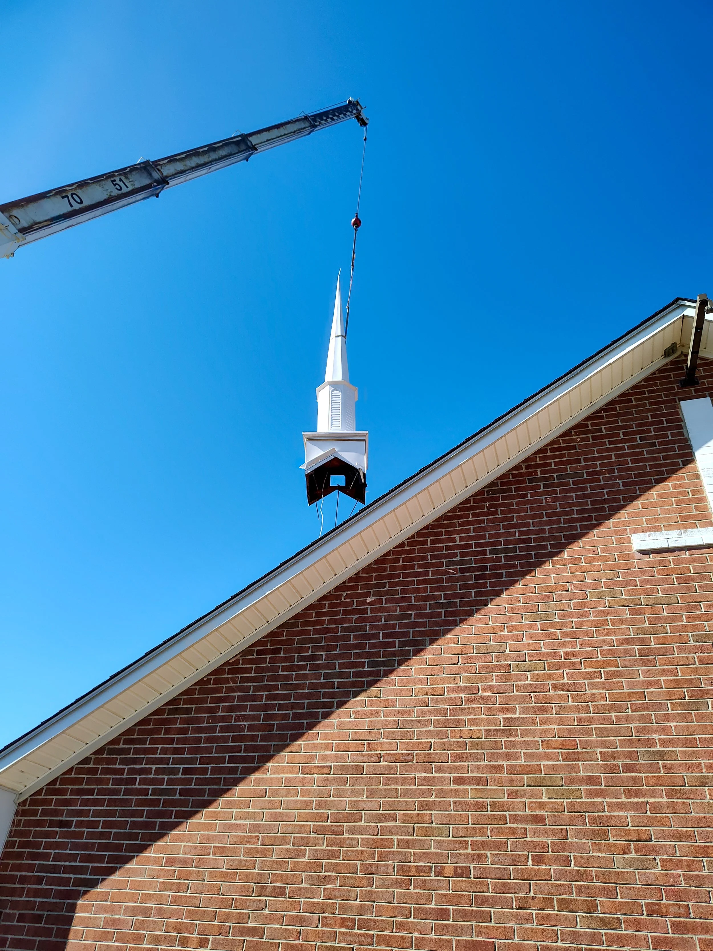 A crane lifts a white steeple onto a red brick church against a bright blue sky.