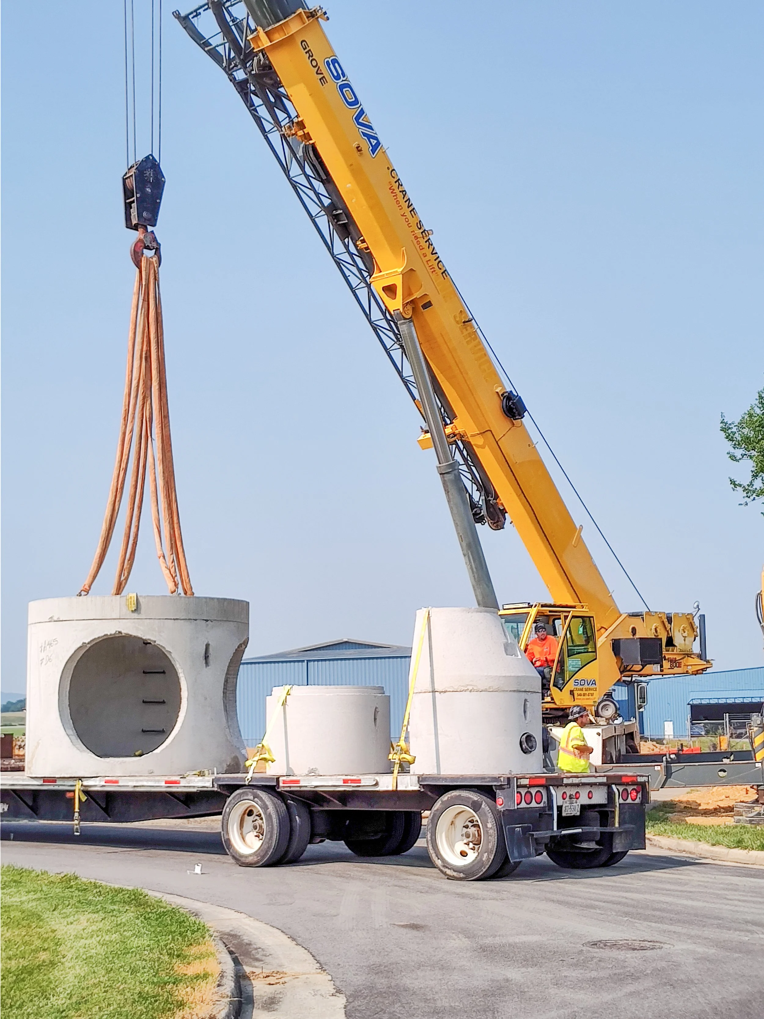 A crane lifting large concrete sewer pipes on a flatbed truck at a construction site with workers nearby.