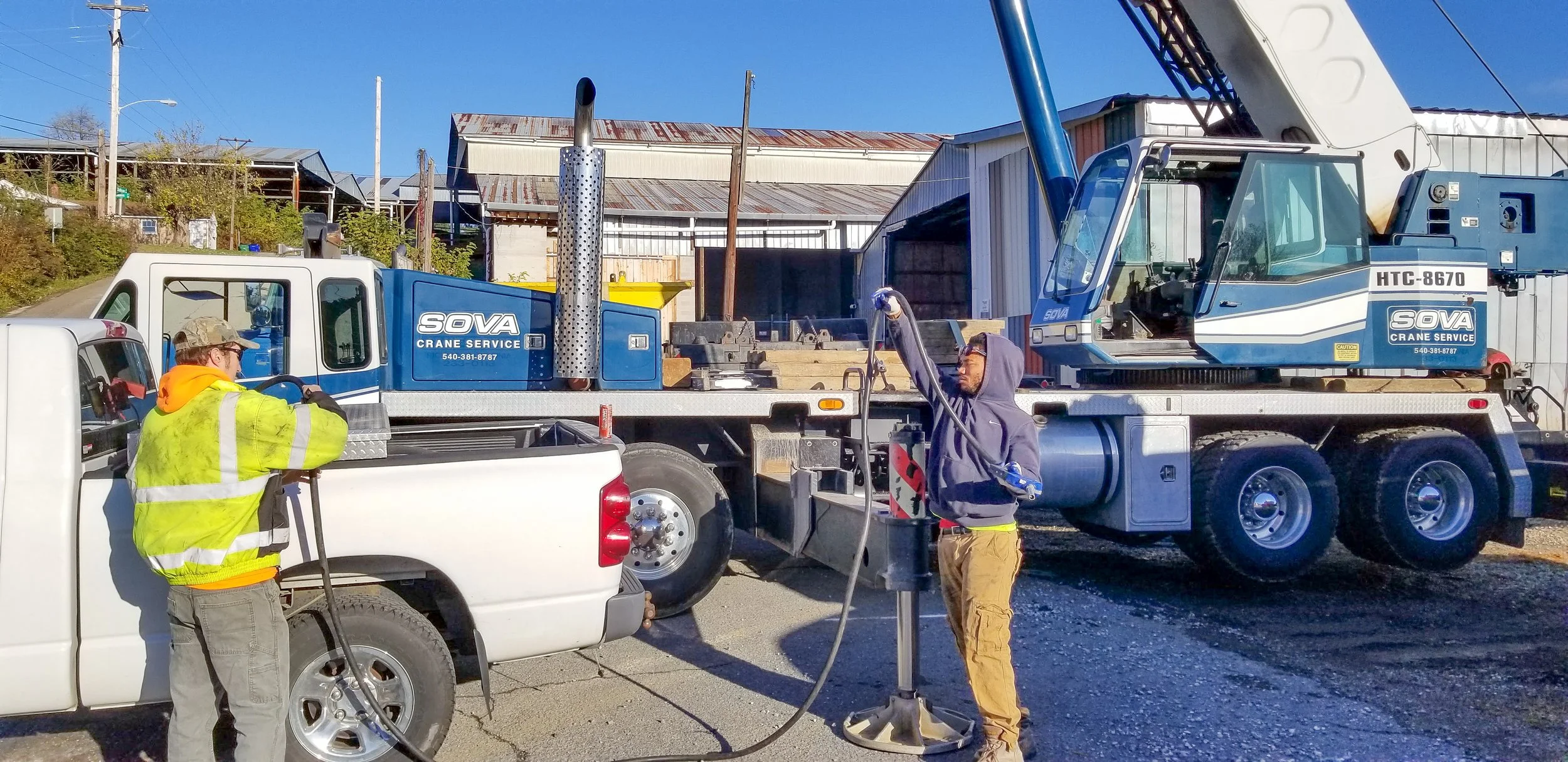Two workers operating a crane service equipment on a flatbed truck, with one man in a yellow safety vest and another in a hoodie, set against a background of industrial buildings and power lines.