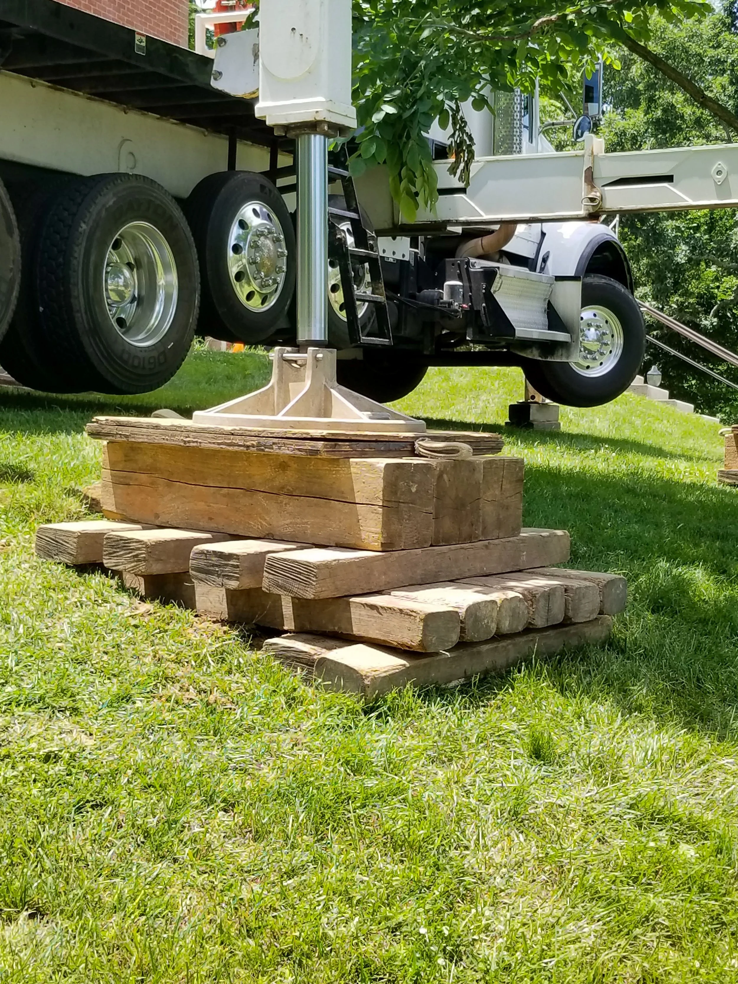 A wooden pile with large logs supports a construction vehicle with black tires and a metal chassis, set on a grassy area under a tree with green leaves.