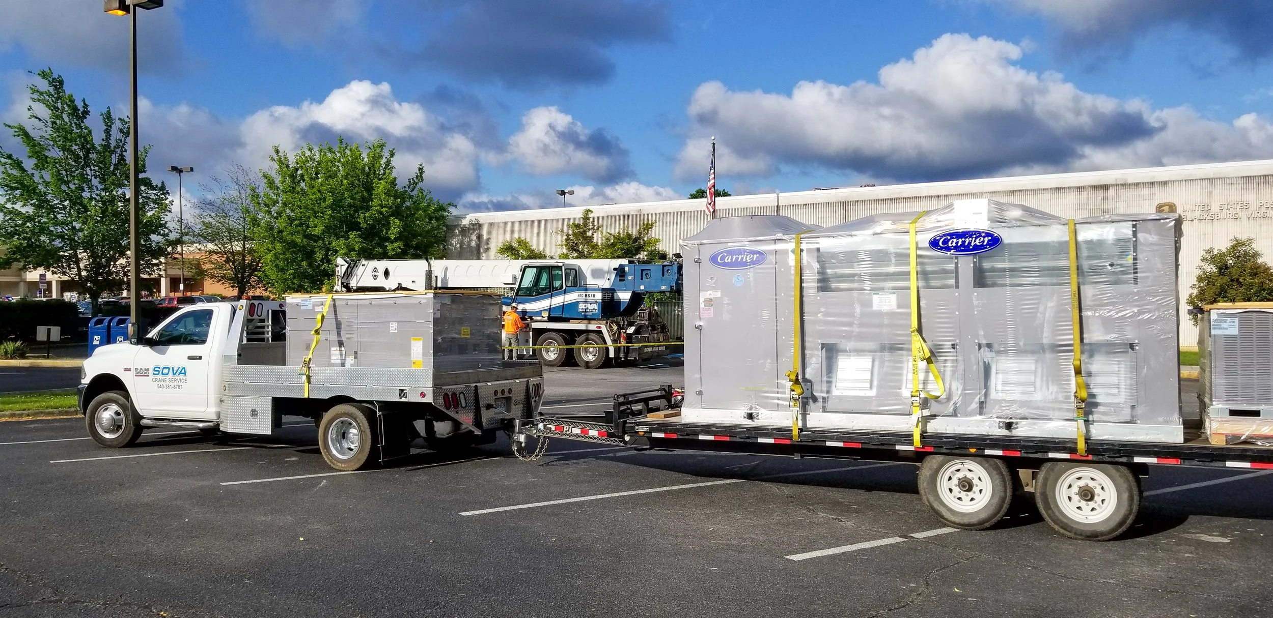 A white truck with a flatbed trailer transporting industrial equipment, including a large metallic object wrapped in plastic with Carrier branding, in a parking lot with trees and a building in the background under a partly cloudy sky.