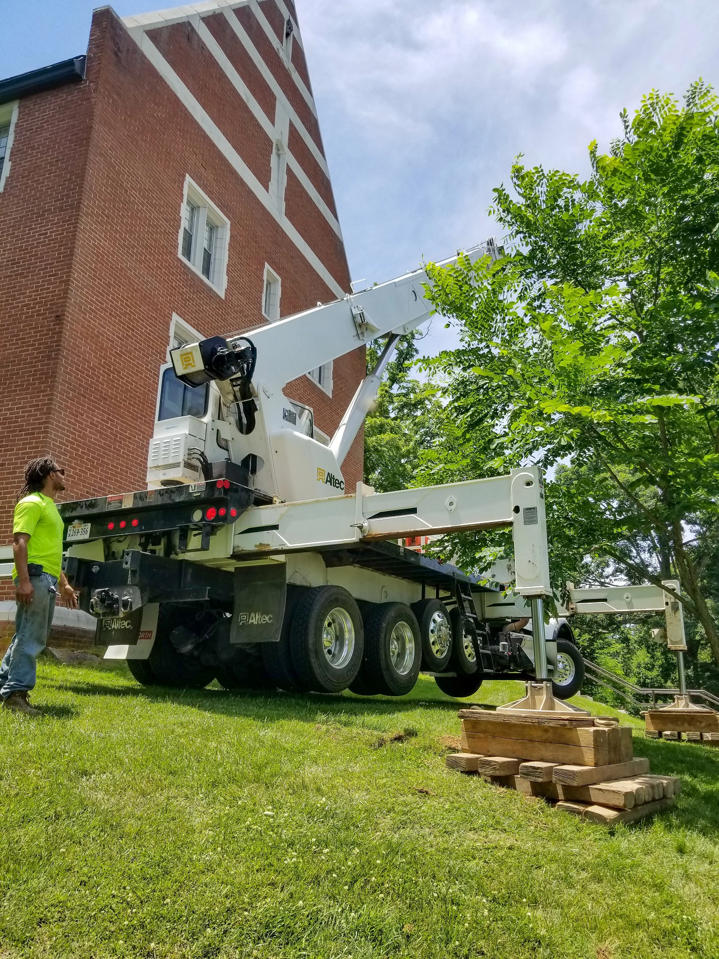 A worker operating a white aerial lift bucket truck near a brick building with trees and a partly cloudy sky.